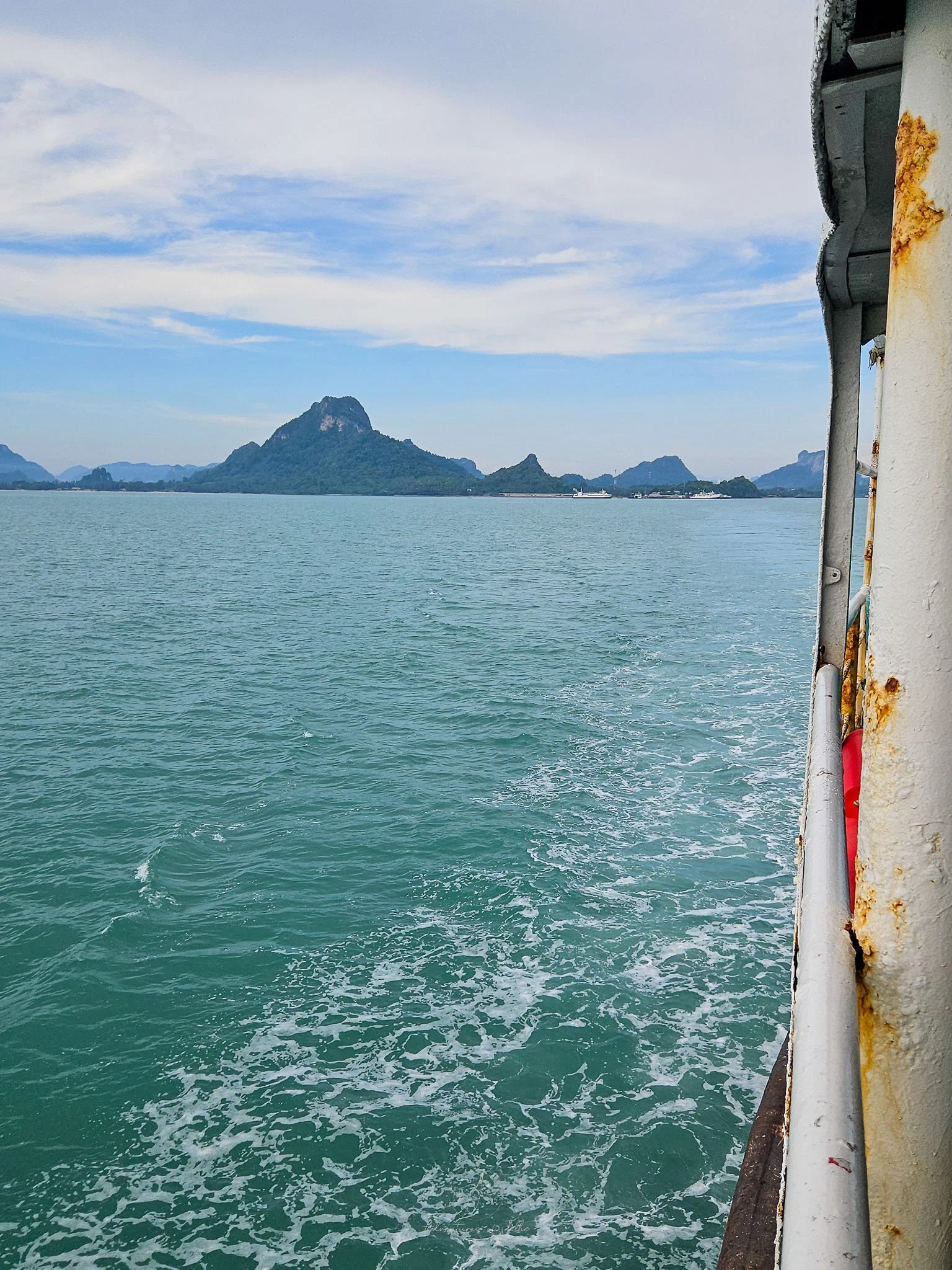 A view from onboard the Raja ferry as it departs from Donsak Pier on the mainland of Thailand. In the distance two white ferries can be seen moored up to the jagged coastline.