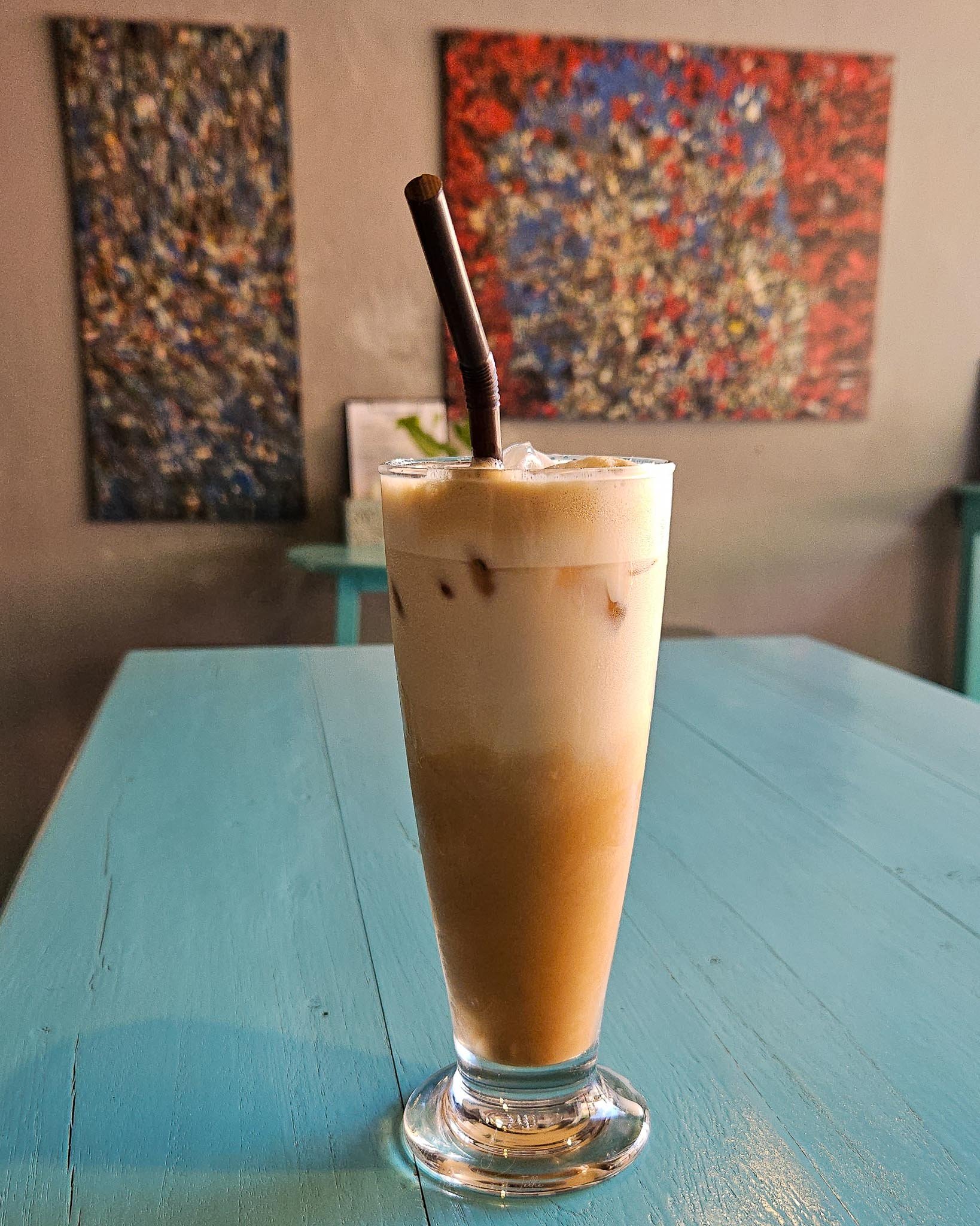 Sitting on a bright turquoise table is a clear glass is filled with iced latte. Behind colourful artwork is displayed on the smooth concrete walls of the Mama Tia's restaurant in Nong Khiaw, Laos.