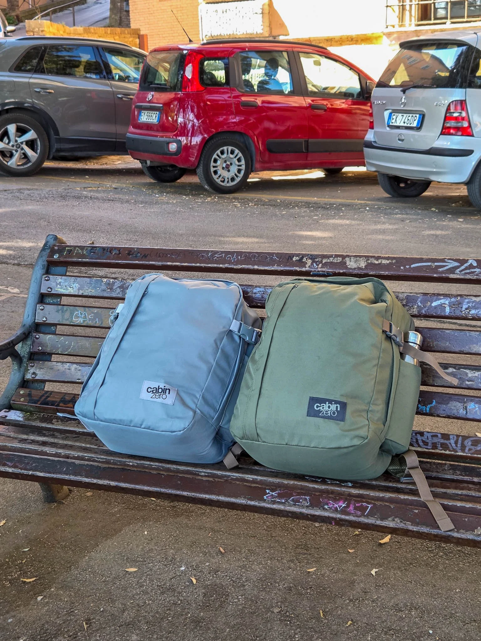Two Cabin Zero Classic Tech 28L bags sits on a wooden bench covered in graffiti. The left hand bag is coloured "Silver Storm" and the right hand bag is the "Georgian Khaki" colour. Behind the bench sit a row of parked cars.