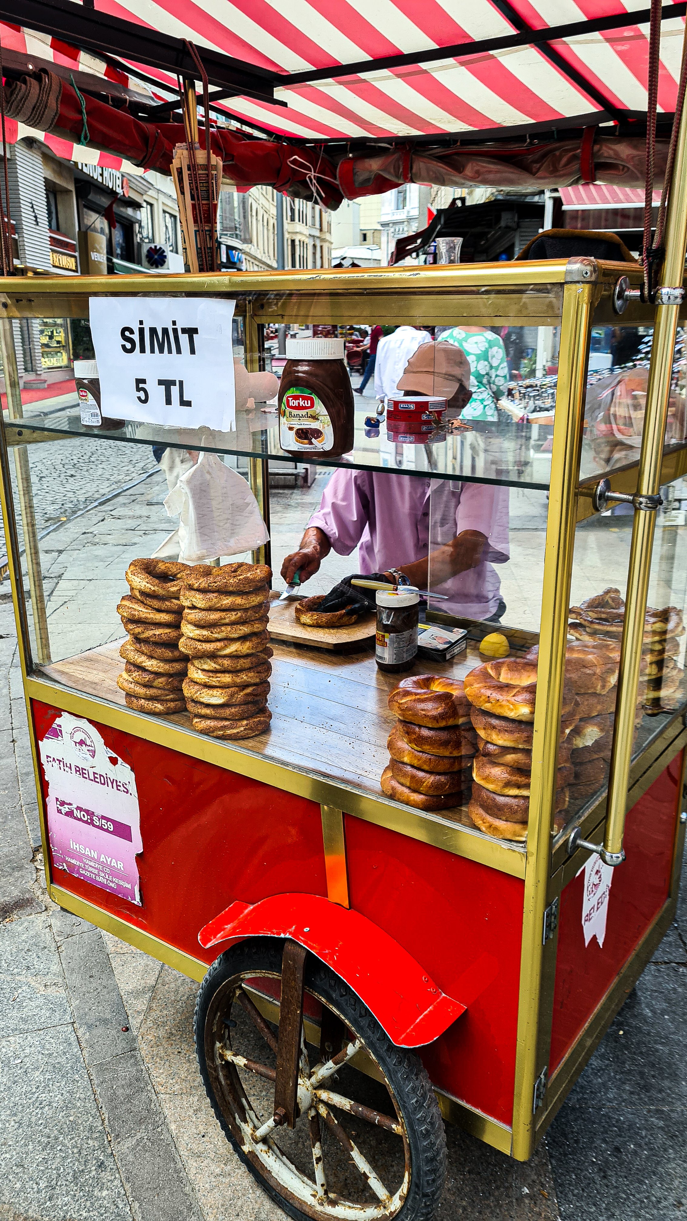 A simit seller stands behind a red cart on the streets of Istanbul. The vendor is seen spreading Nutella onto a sliced simit.