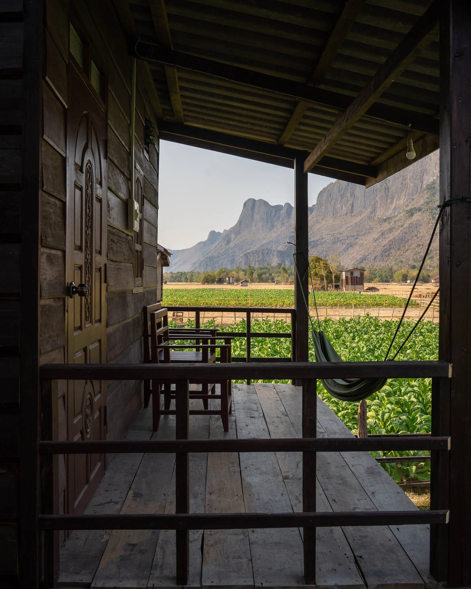 A hammock and wooden chairs sit on a balcony of one of the Kong Lor Eco Lodge Bungalows. Behind tobacco plantations stretch away to mountains.