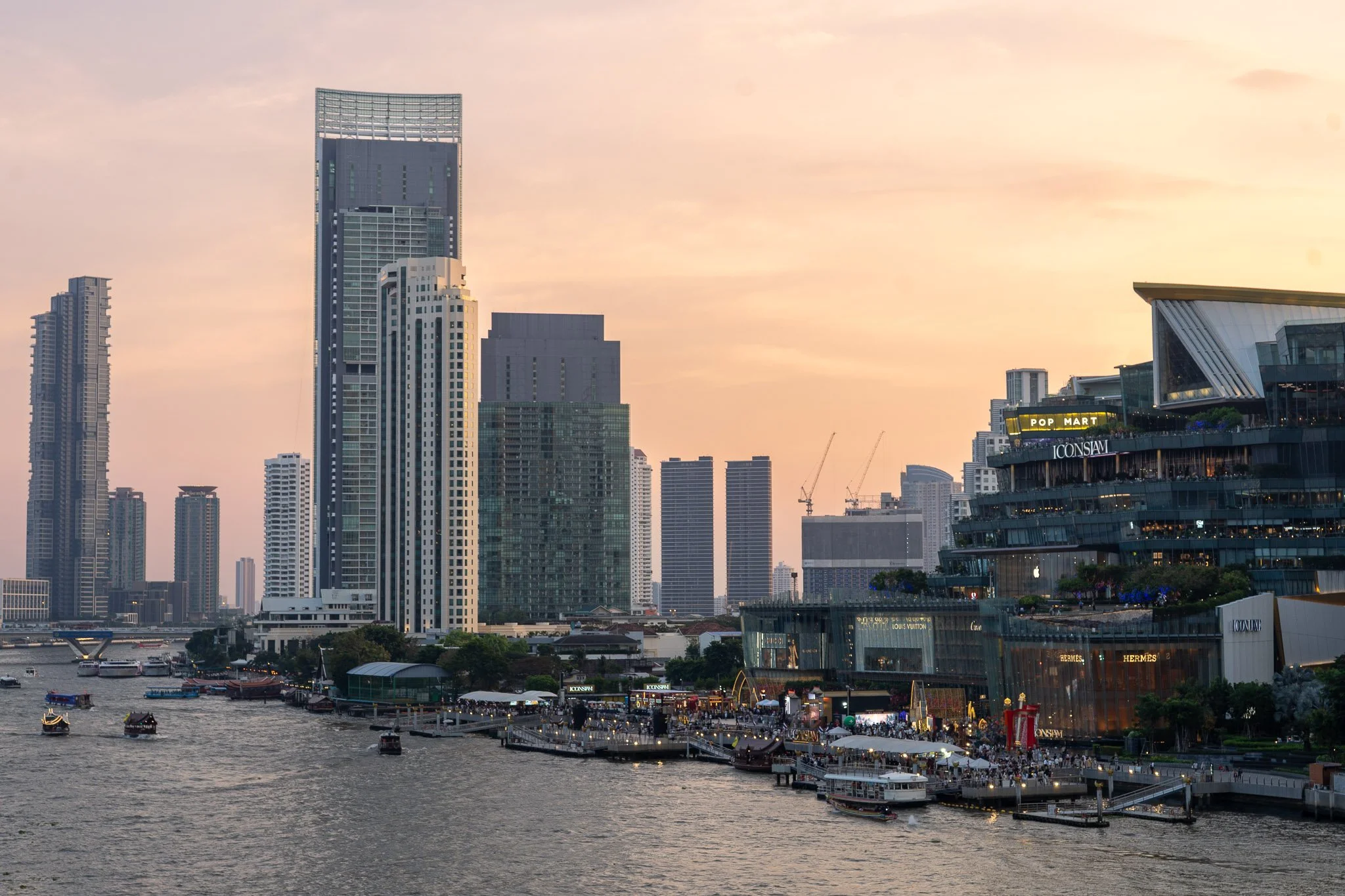 The sun sets behind Bangkok's iconic Icon Siam shopping mall. The sky is lit up orange behind the sky scrapers and high-end hotels that line the Chao Phraya River.