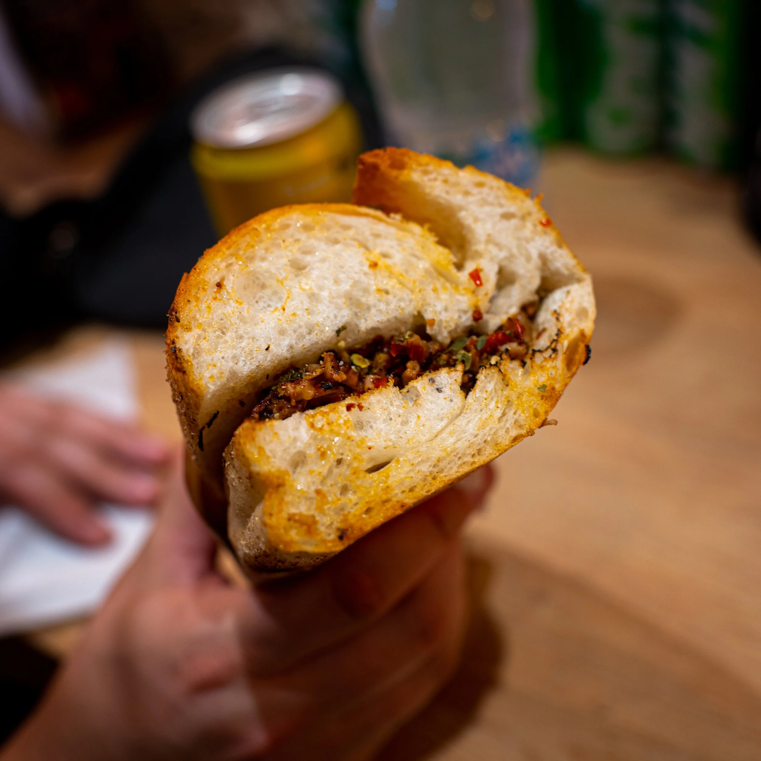 A  toasted kokorec sandwich is held up to the camera. The kokorec is stuffed full of lambs offal, chilli and chopped onions.