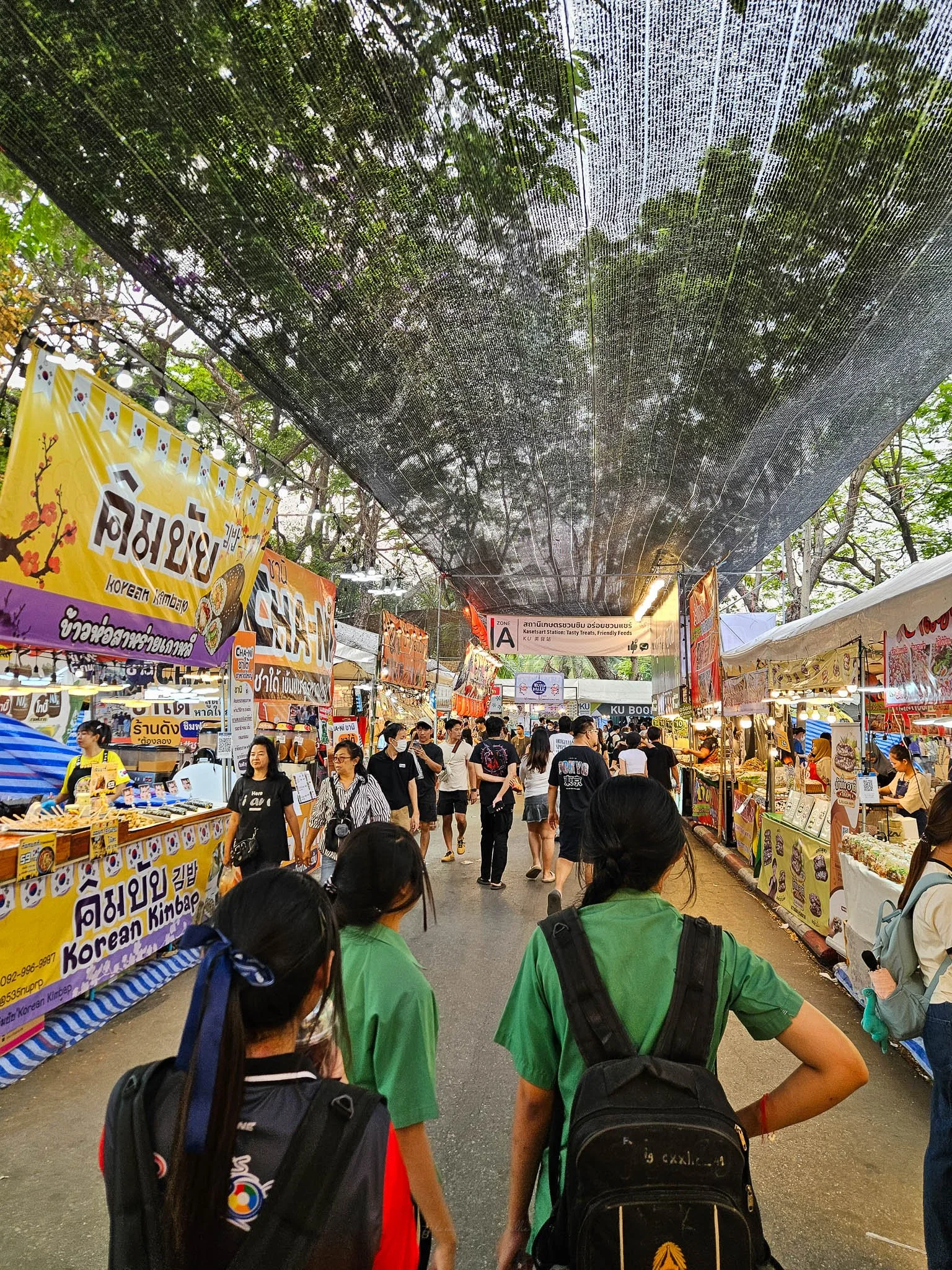Crowds of people walk through the Kaset Fair Market. On either side, street food stalls line the road selling everything from Korean Bimbap to Thai style salads. In front of the viewer three Thai school girls wearing backpacks walk in front.