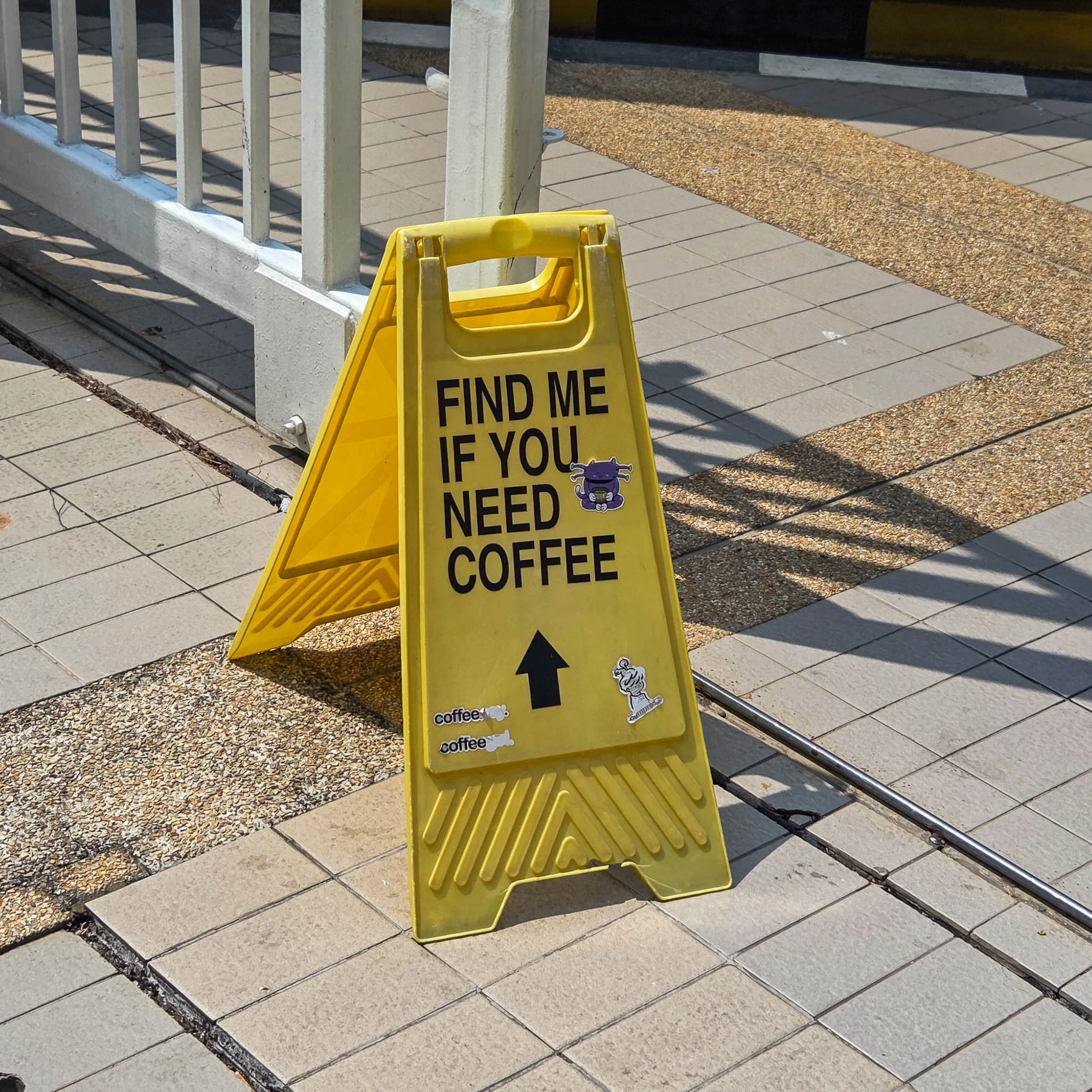 A plastic yellow cleaning sign has the words "Find me if you need coffee" stuck on it in black capital letters. At the bottom is a black arrow pointing up and also a purple cat sticker.
