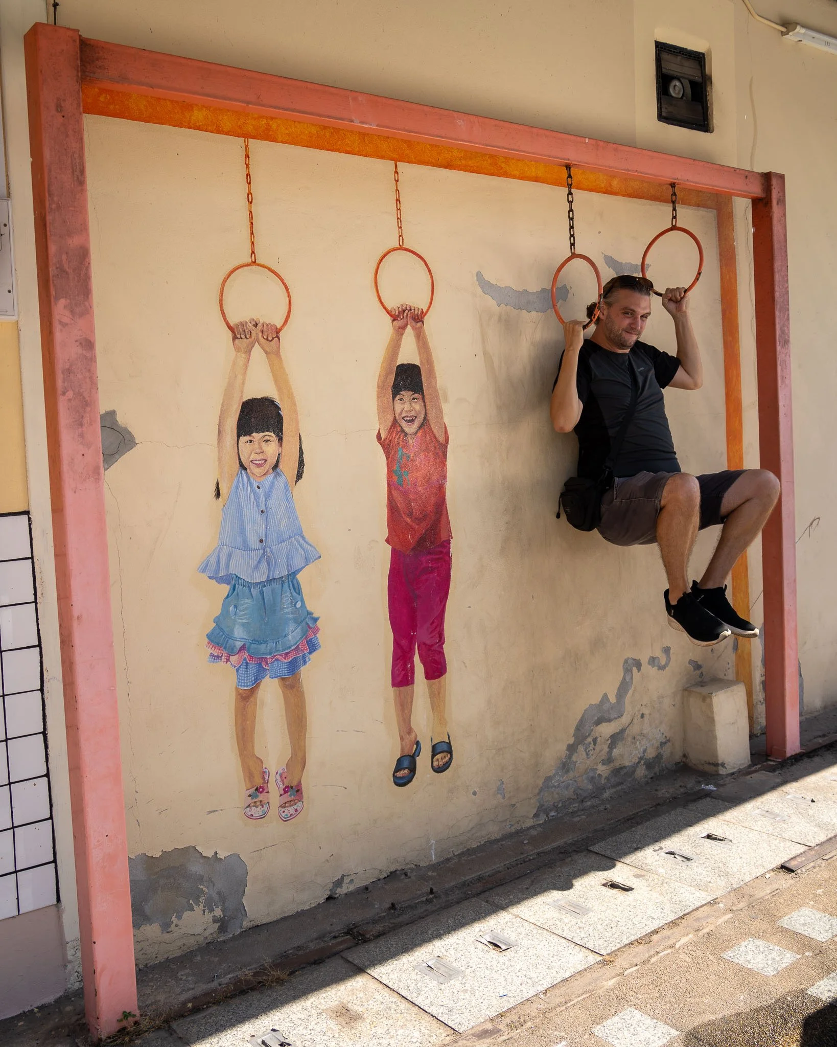 John hangs from monkey bars at the Khon Kaen Art Wall. Next to him are two girls painted onto the wall also hanging from the monkey bars.