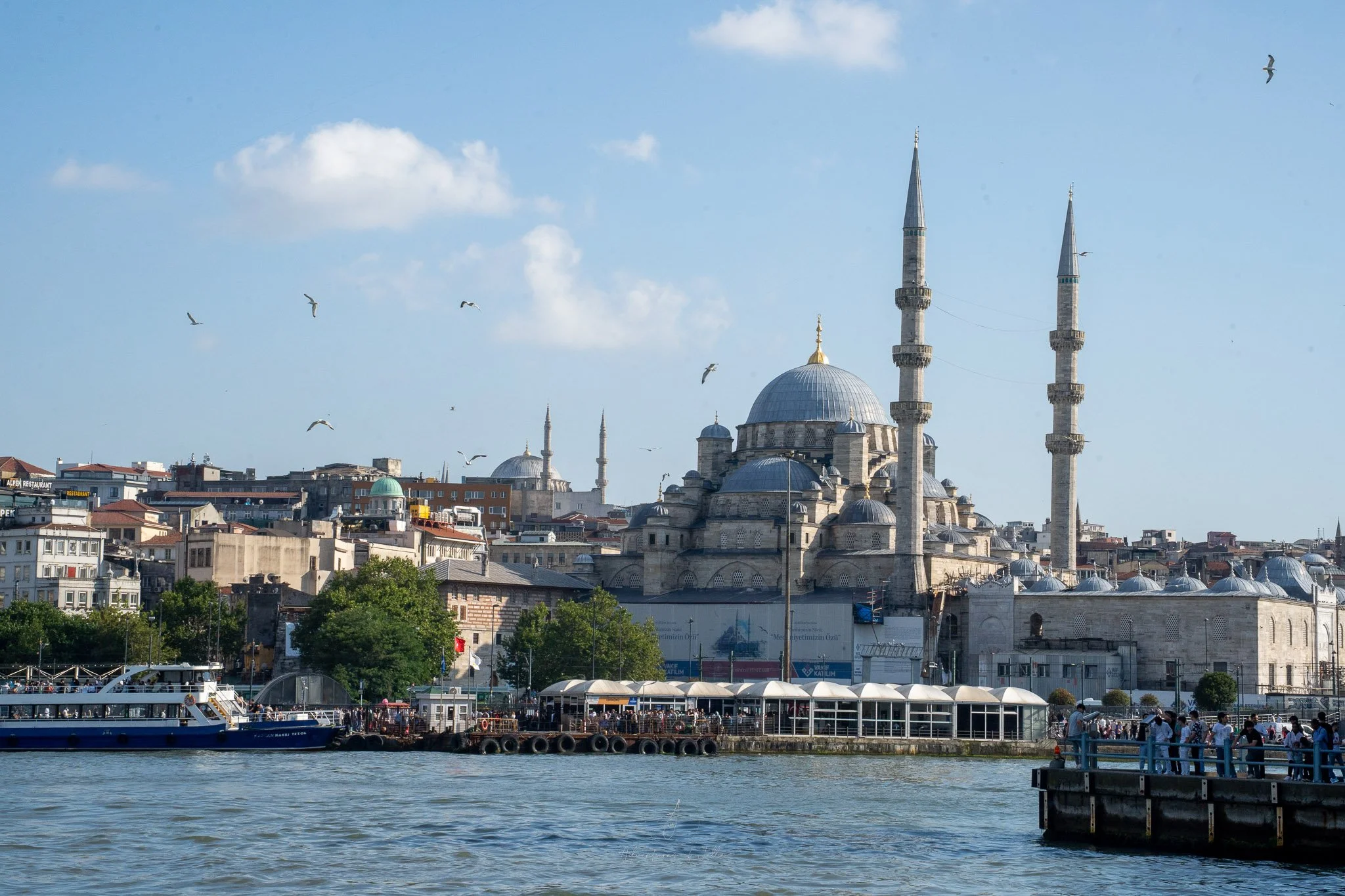 The skyline of Istanbul rises up above the waterfront. On the shoreline people crowd for photos. In front the domed towers and minarets of the Blue Mosque rise up to blue sky.