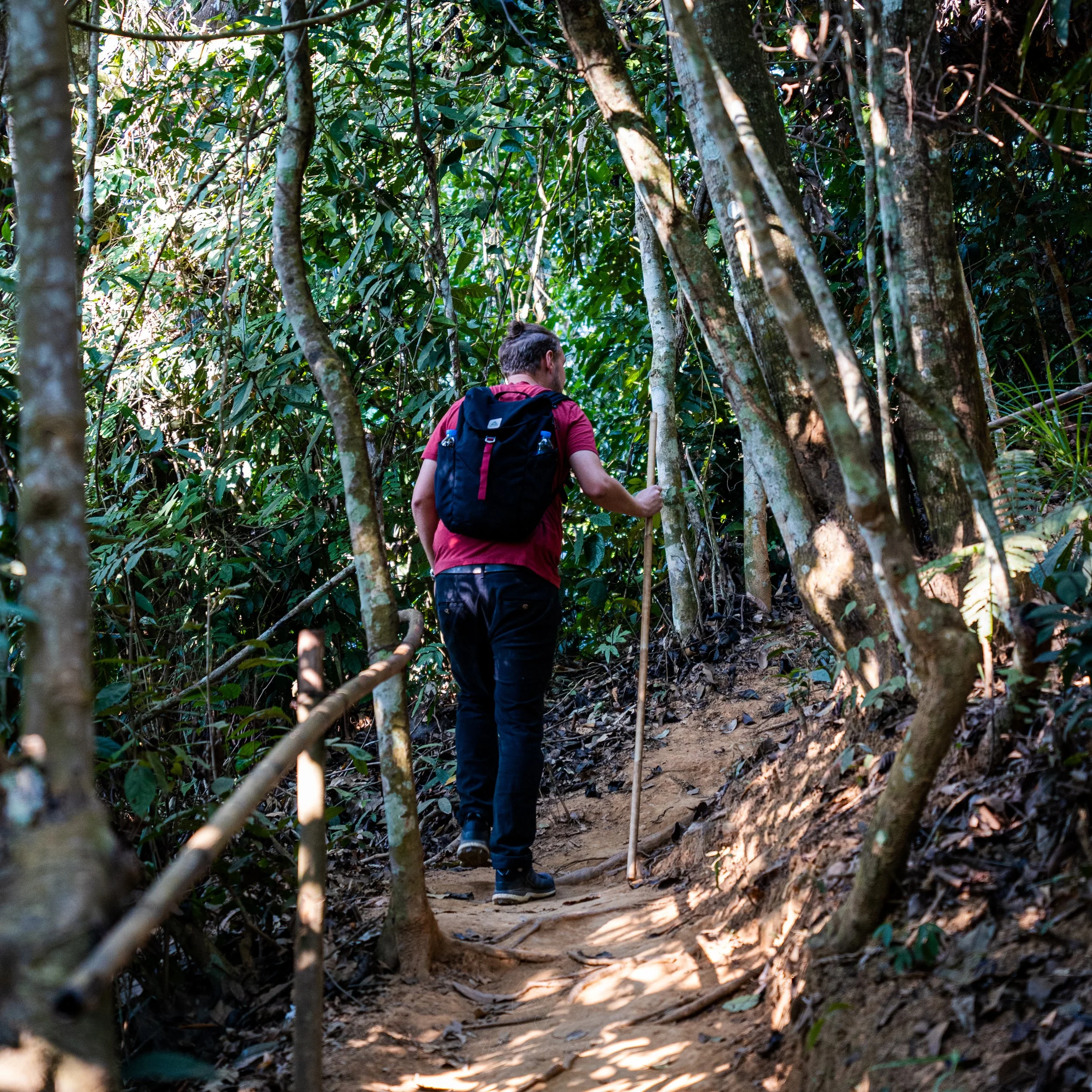 John hikes with a stick up a dusty, forested path to the top of the Som Nang Viewpoint. He is wearing a red t-shirt, black trousers and a Salkan daypack.