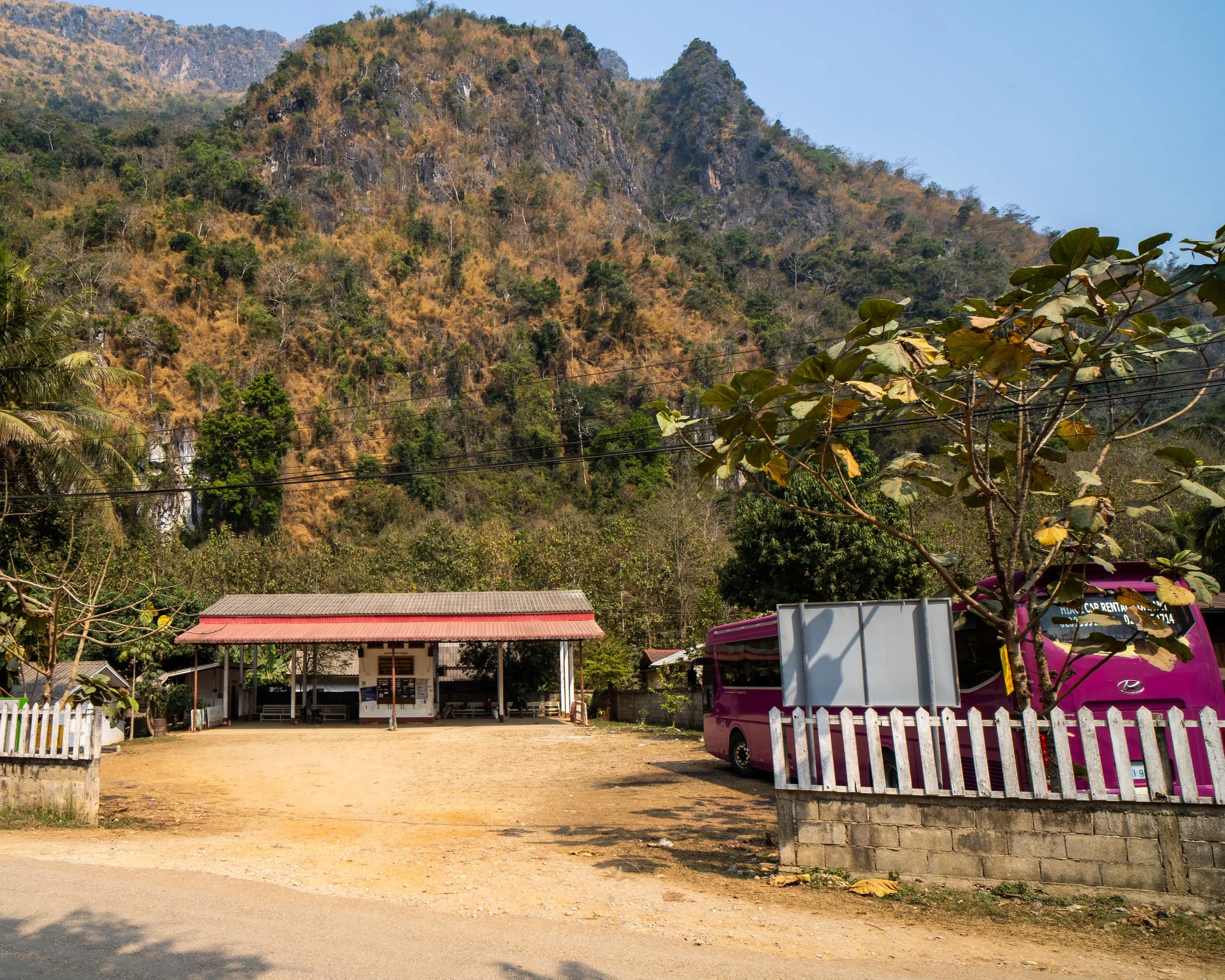 The entrance to Nong Khiaw’s Bus Station. To the right a purple coach sits parked in the dusty car park. In front sits the red-roofed ticket office.