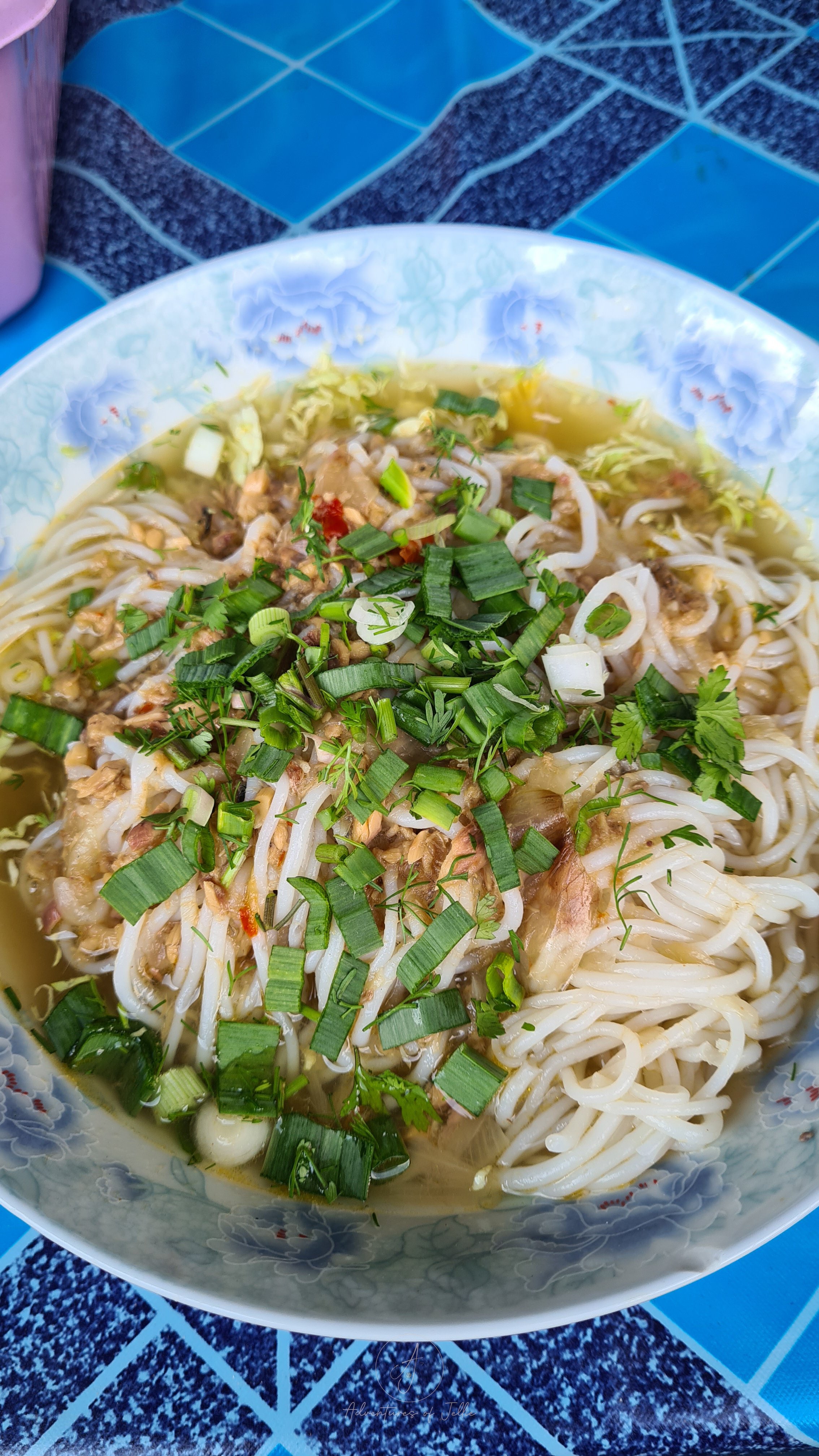 A pale blue bowl is filled with fish noodle soup which includes flakes of brown fish meat, rice noodles, beansprouts and slices of spring onion. The bowl sits on a blue table in a street side restaurant in Nong Khiaw, Laos.