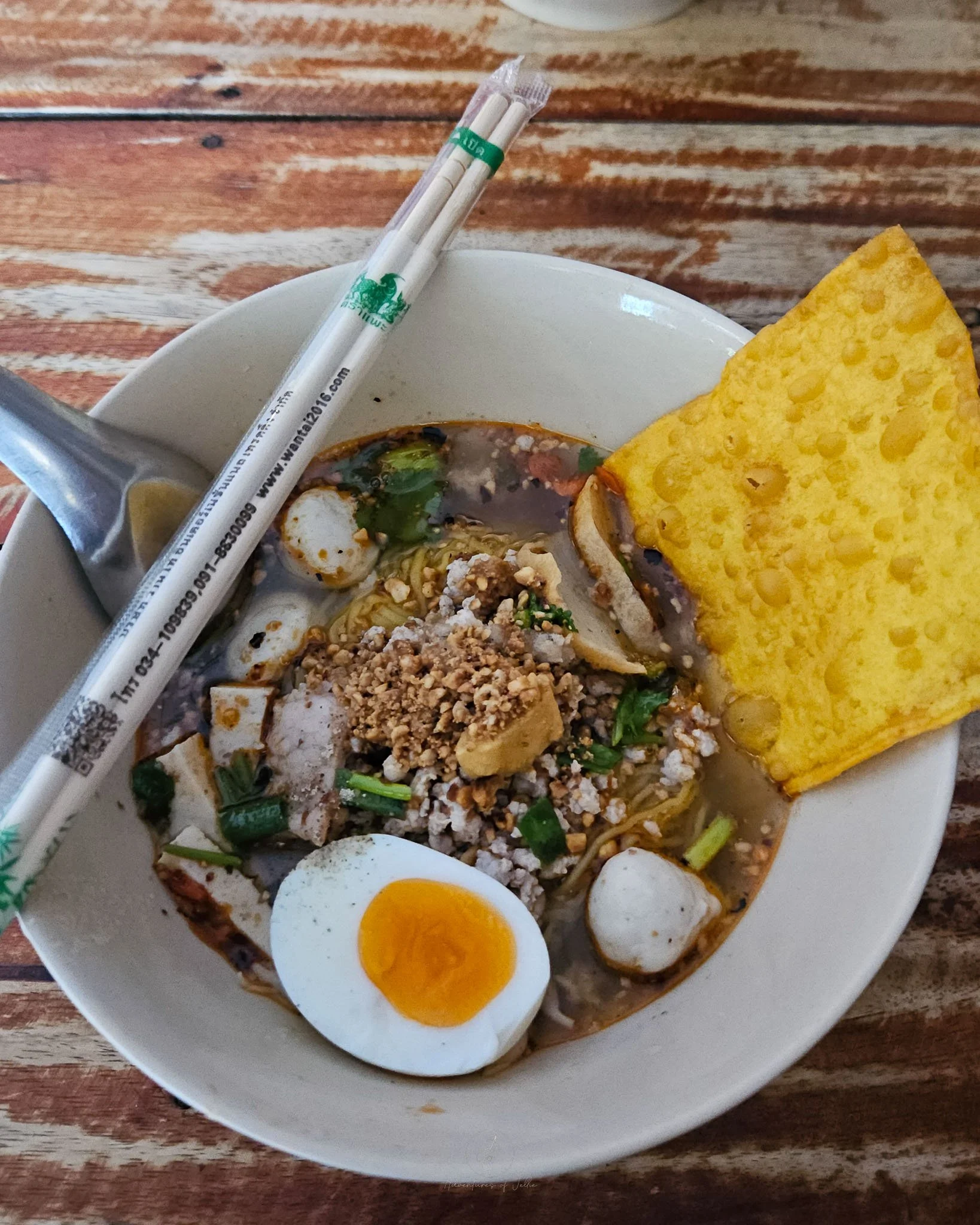 In a china bowl sits tom yum noodle soup. The egg noodles and soup are topped with pork balls, slices of pork, pork sausage, half a boiled egg, a wonton crisp and chop sticks.