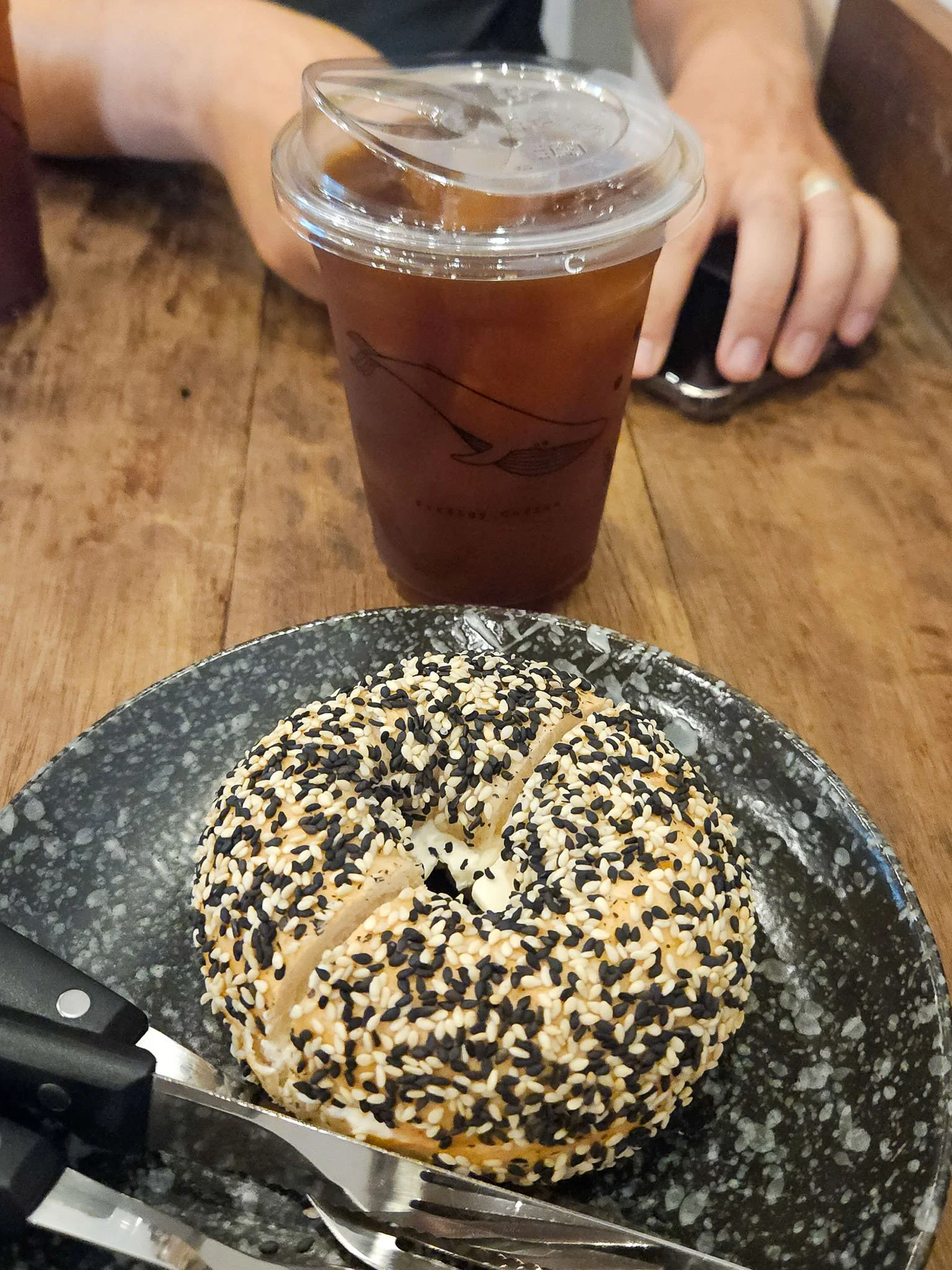A cup of iced americano sits next to a sesame covered cream cheese bagel sitting on a dark grey plate at the Prodigy Coffee shop in Khon Kaen, Isan.
