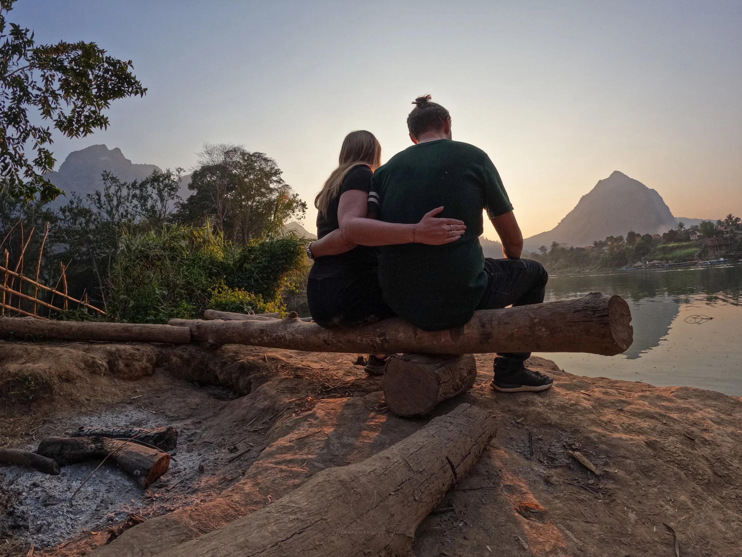Adventures of Jellie sit arm in arm watching the sunset from a log bench overlooking the Nam Ou River in Nong Khiaw, Laos.