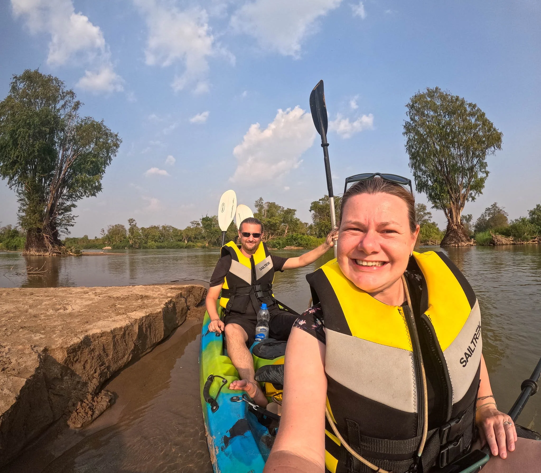 Adventures of Jellie pose for a GoPro selfie whilst sat in a blue kayak. They are both wearing a yellow and black lifejacket and John is holding up a black oar in his left hand.