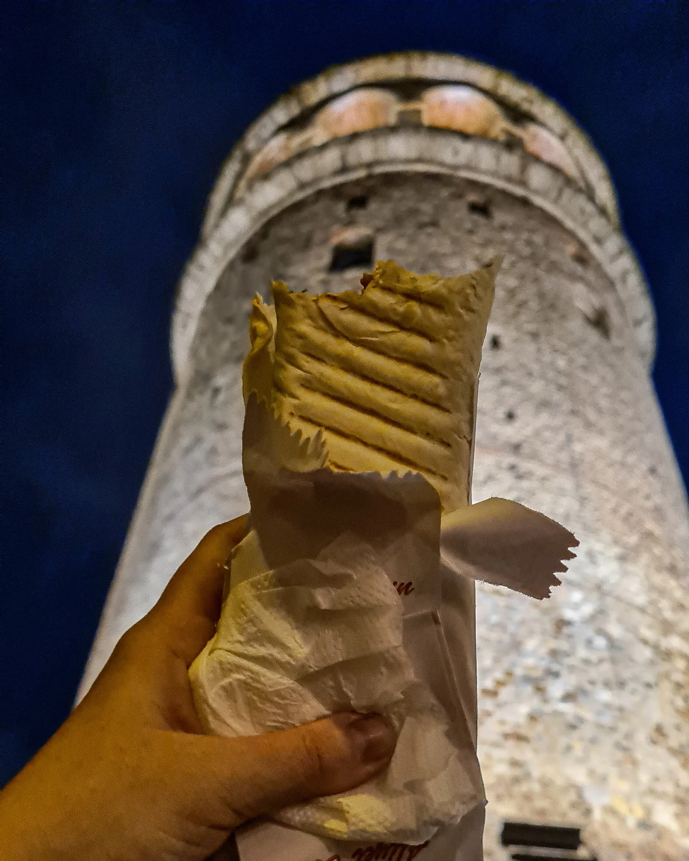 A doner kebab sits inside a white paper bag and is held up to the camera. Behind the Galata Tower rises up above the view. Photo take in Istanbul at night.