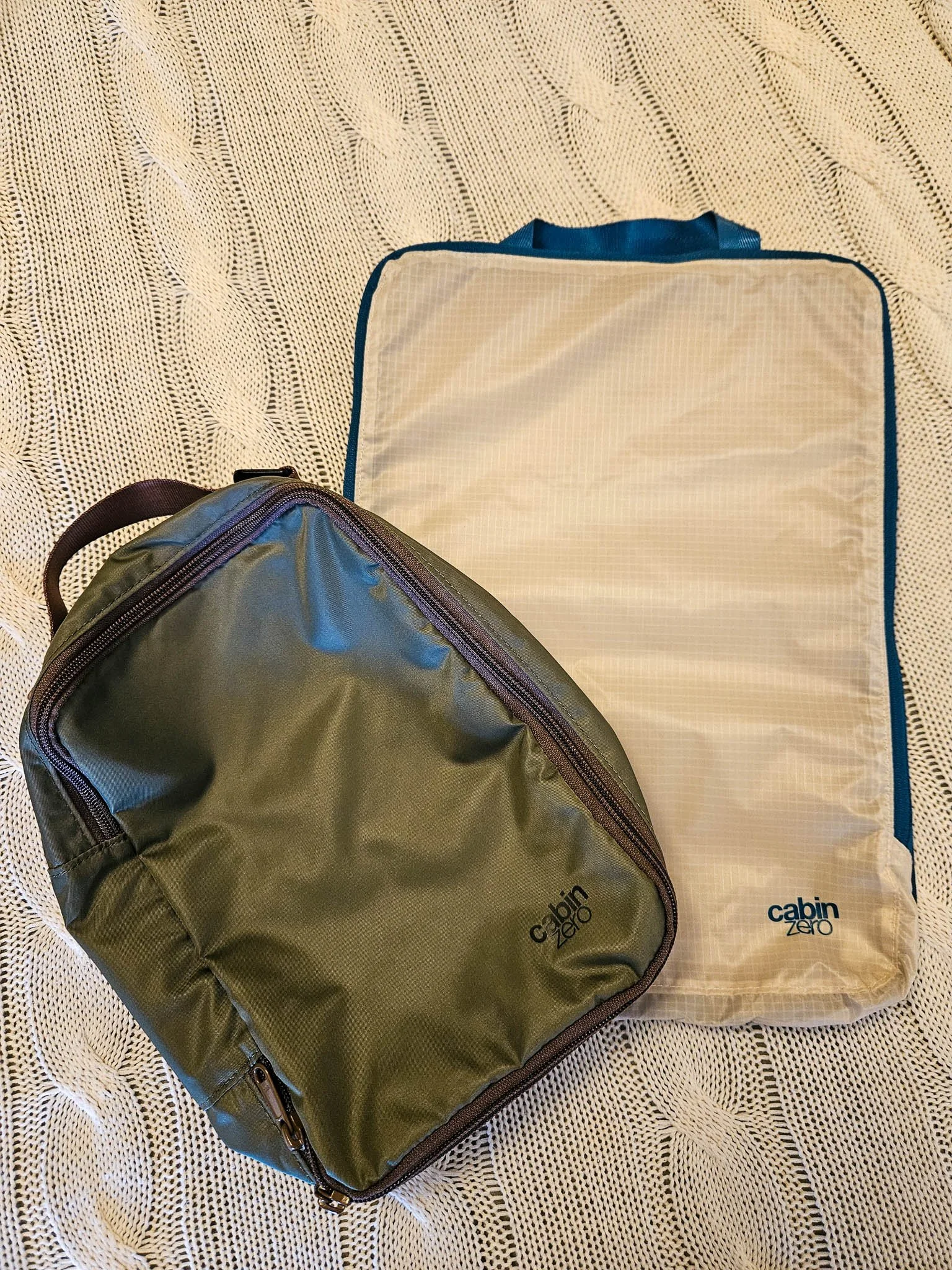 On a bed sit a medium, khaki green coloured Cabin Zero lux compression packing cube and next to it, is a white large ultralight packing cubes.