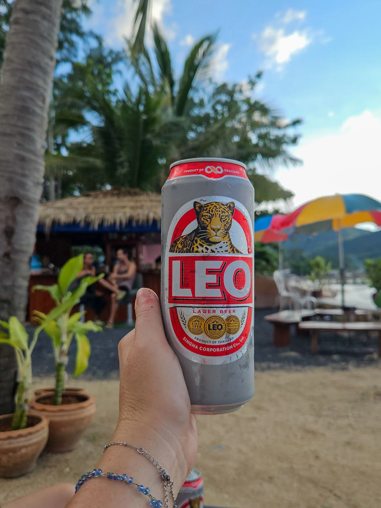 A can of chilled Leo beer is held up to the camera at a beach bar on the shoreline of Chaloklum Beach.