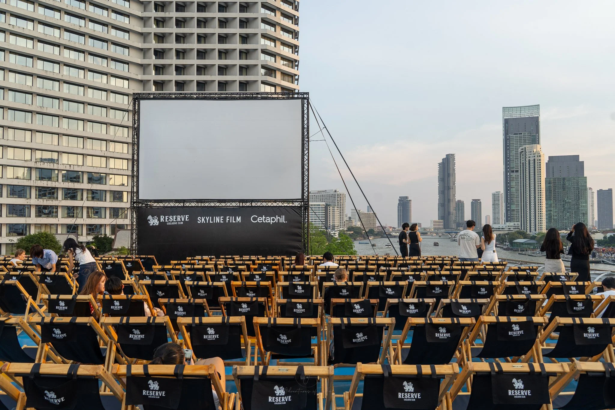 Black wooden deckchairs sit in horizontal rows in front of a cinema screen on top of the Rivercity rooftop in Bangkok. Behind the screen the Sheraton Hotel rises up and to the right of this, the Chao Phraya river stretches away.
