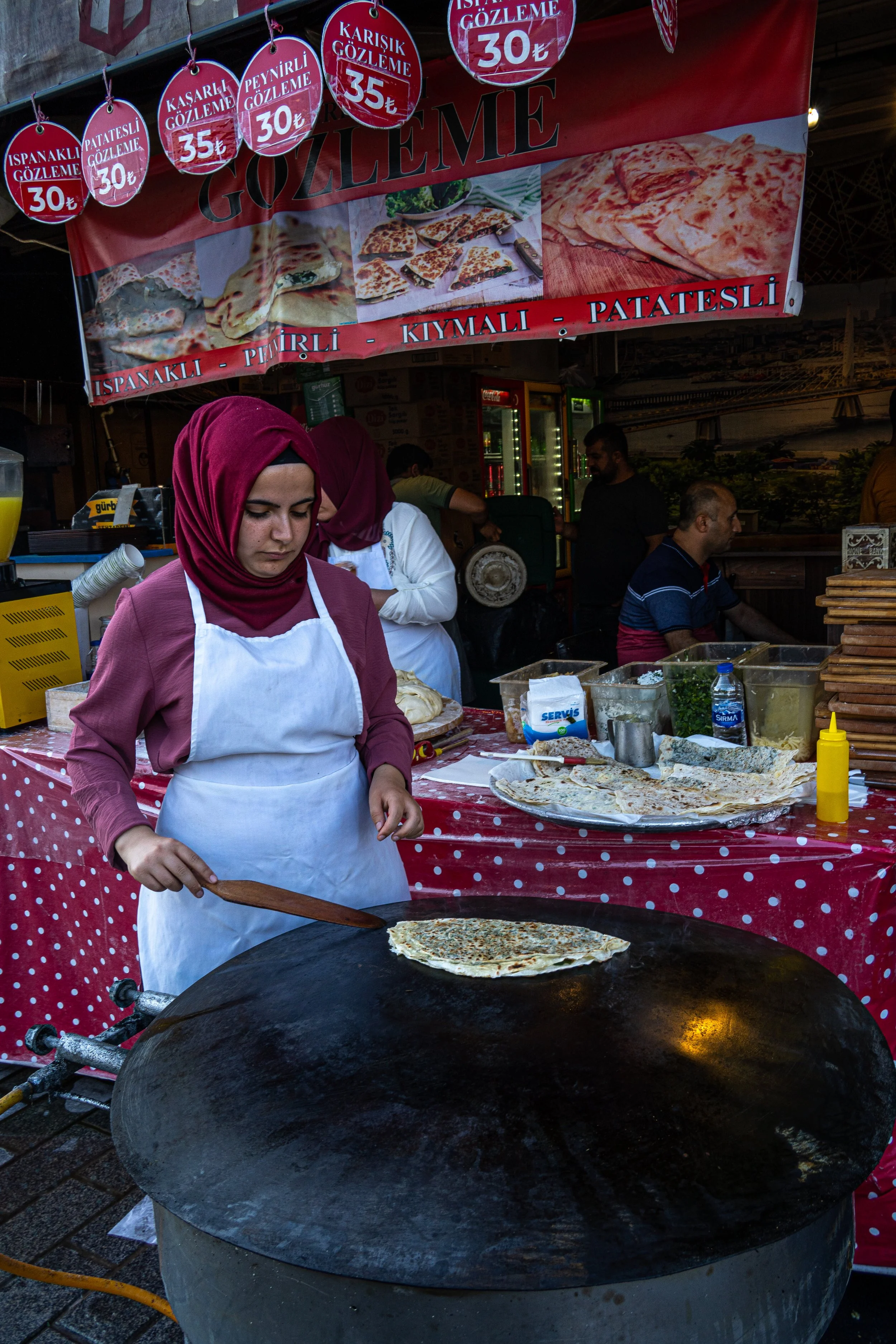 A lady wearing maroon clothes stands frying a gözleme on a large frying pan on the streets of Istanbul.