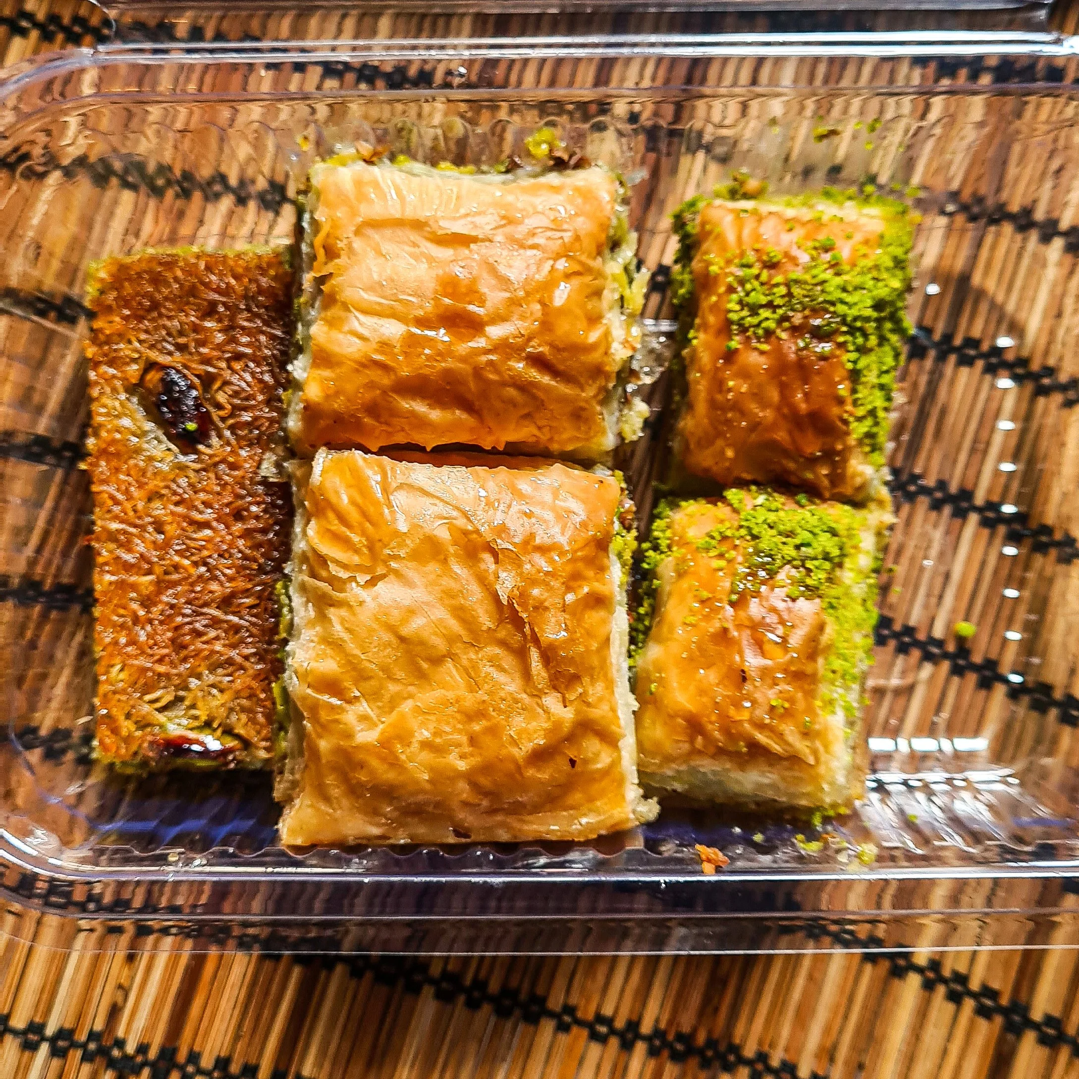 A clear plastic box is filled with different kinds of baklava. Including two squares covered in pistachio, two stuffed with walnuts and one covered in shards of pastry and dried fruit.
