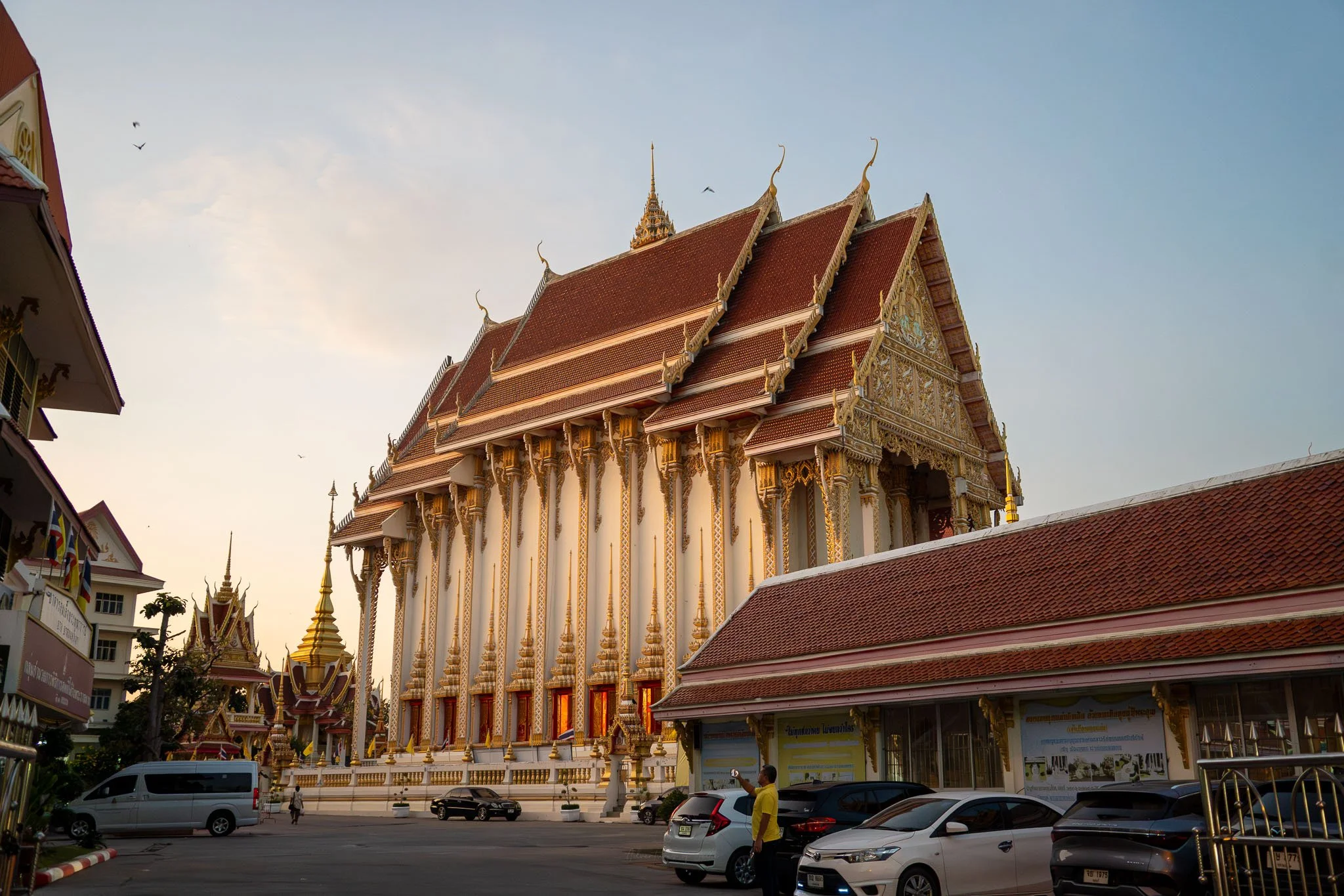 The enormous white, gold and red prayer hall rises up in front of the viewer at the entrance to A pigeon flies away from the golden stupa of Wat That Phra Aram Luang Temple in Khon Kaen. In the foreground a man wearing yellow takes a photo.