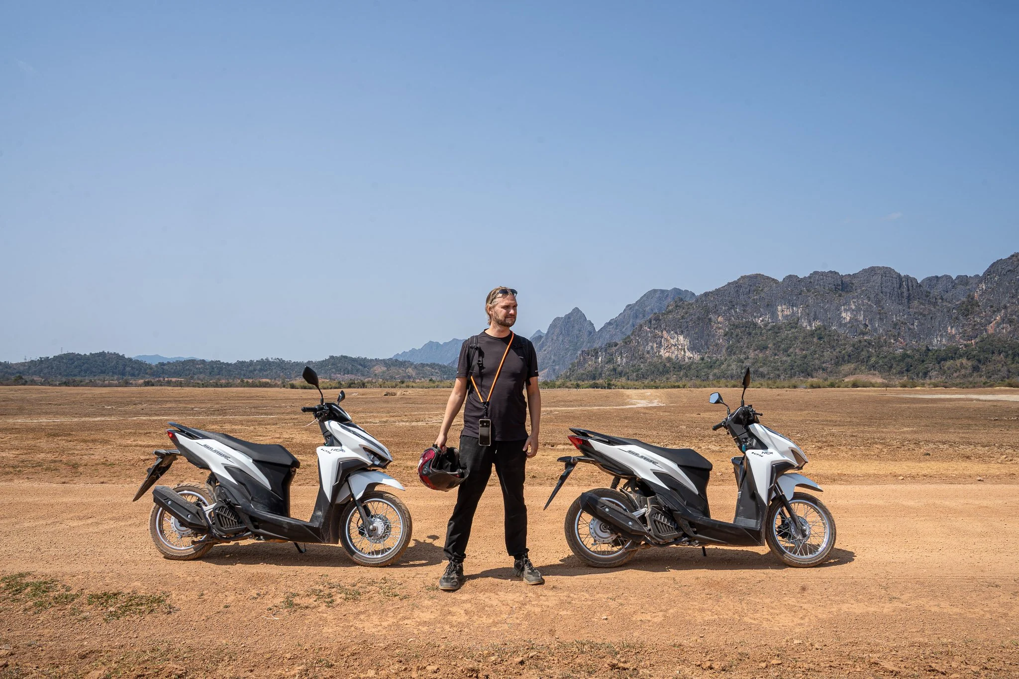John stands in the middle of two white Honda Click scooters. He is looking to the left, wearing a black t-shirt, trousers and trainers whilst holding a red motorbike helmet in his hand. Behind jagged limestone karsts and sandy earth stretch away.