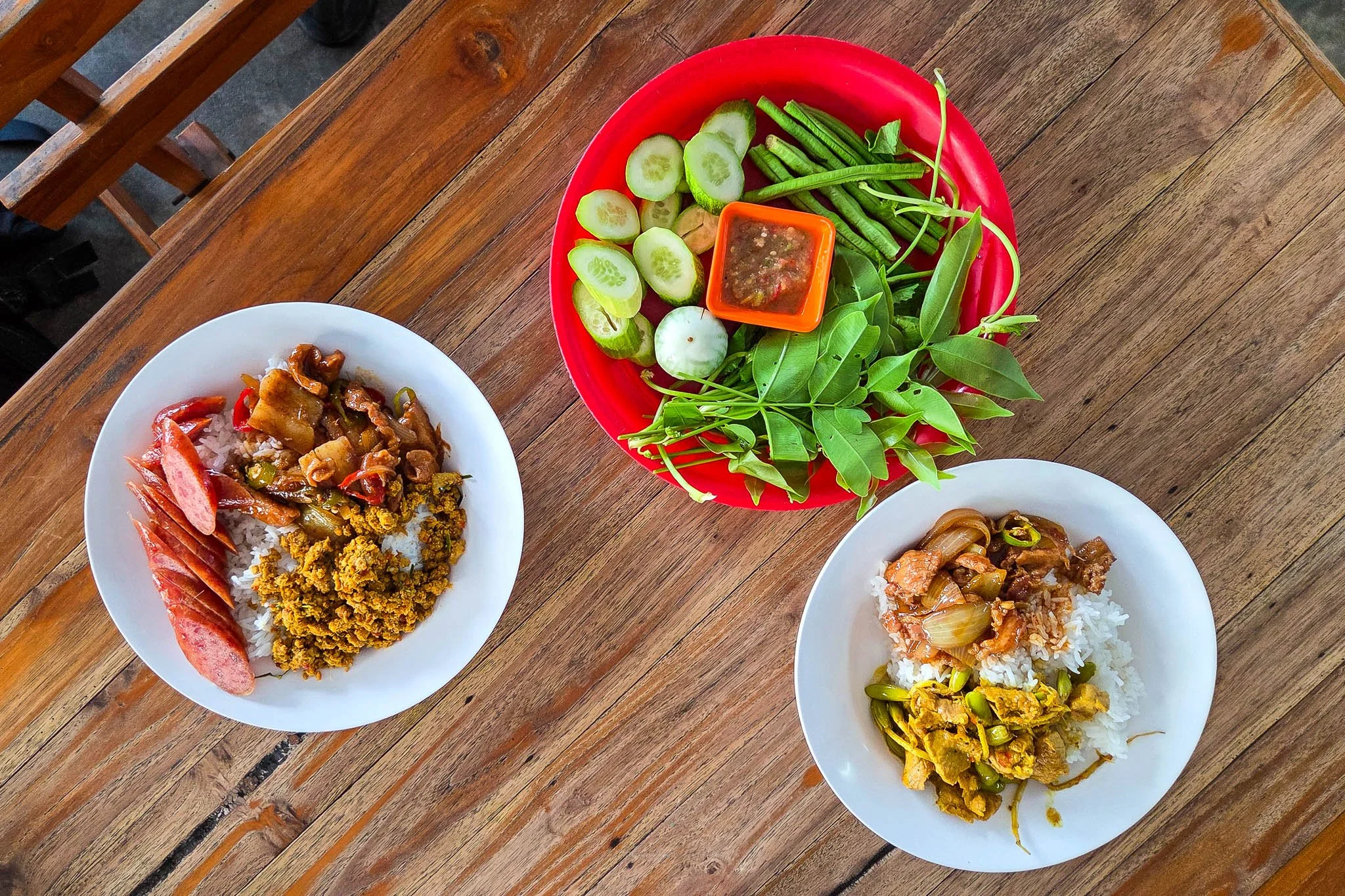 A wooden table is filled with three plates. Two of the plates are white in colour and are filled with rice, curries and Chinese sausage. The third plate is bright red and is full of raw green vegetables and a dipping sauce.