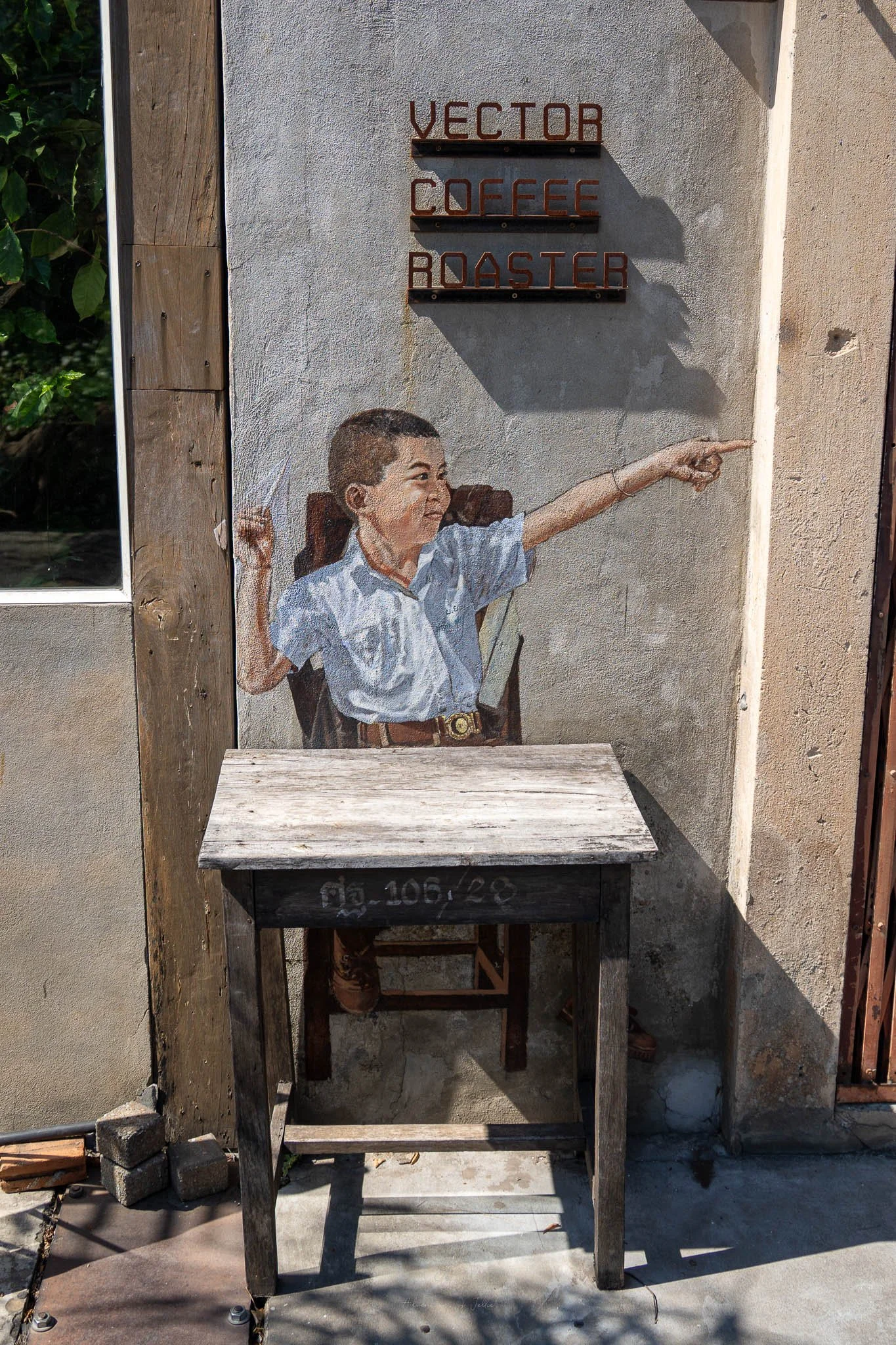 A school boy is painted on the side of a wall in Khon Kaen. The boy is wearing a blue shirt and is holding a paper aeroplane in his right hand. The boy sits behind a wooden desk beneath a sign for Vector Coffee Roaster.