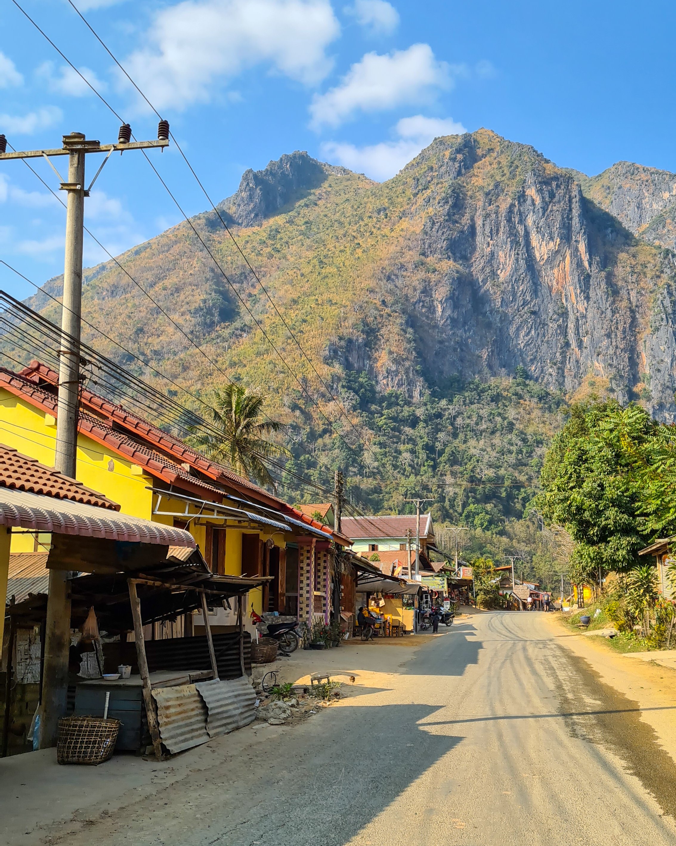 The main road running through the town of Nong Khiaw in Laos is lined with small shops, bars, restaurants and tour agencies. In the distance a huge limestone mountain towers over the viewer.