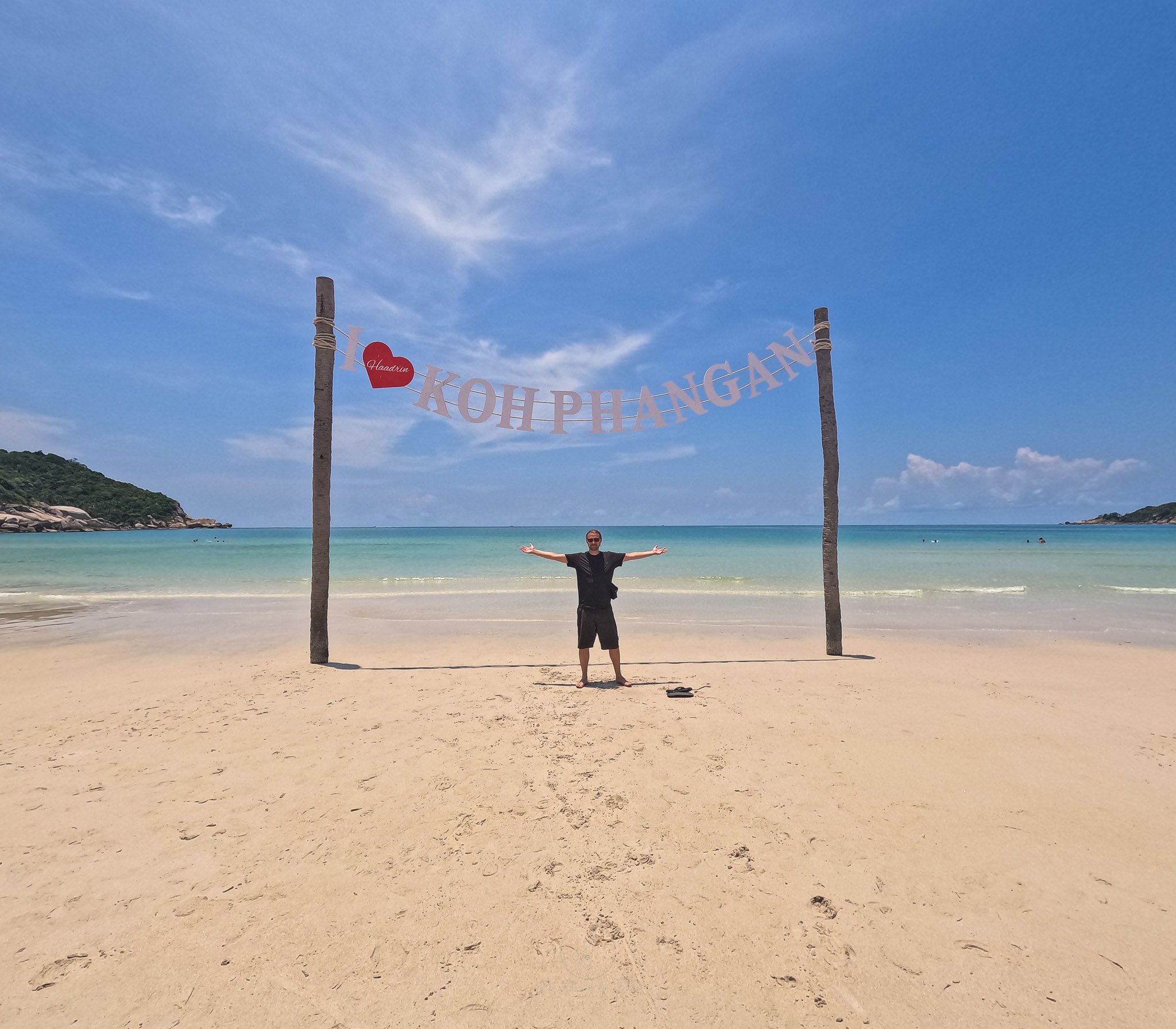 John stands wearing a black t-shirt and shorts with his hands outstretched underneath a sign which reads "I heart Koh Phangan".