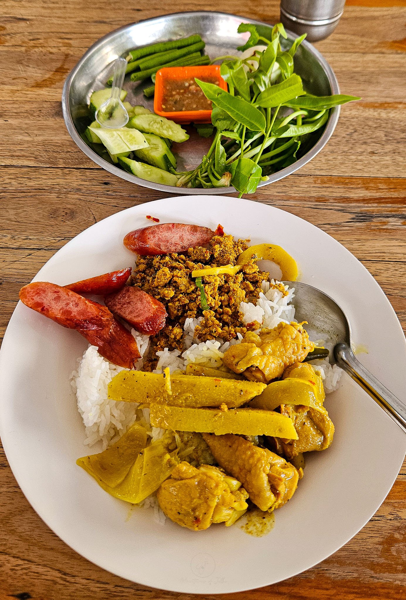 In the foreground a white plastic plate is filled with a yellow chicken and bamboo shoot curry alongside a mince beef kra pao and Chinese sausage all sitting on top of white rice. Above sits a metal plate filled with raw green vegetables.