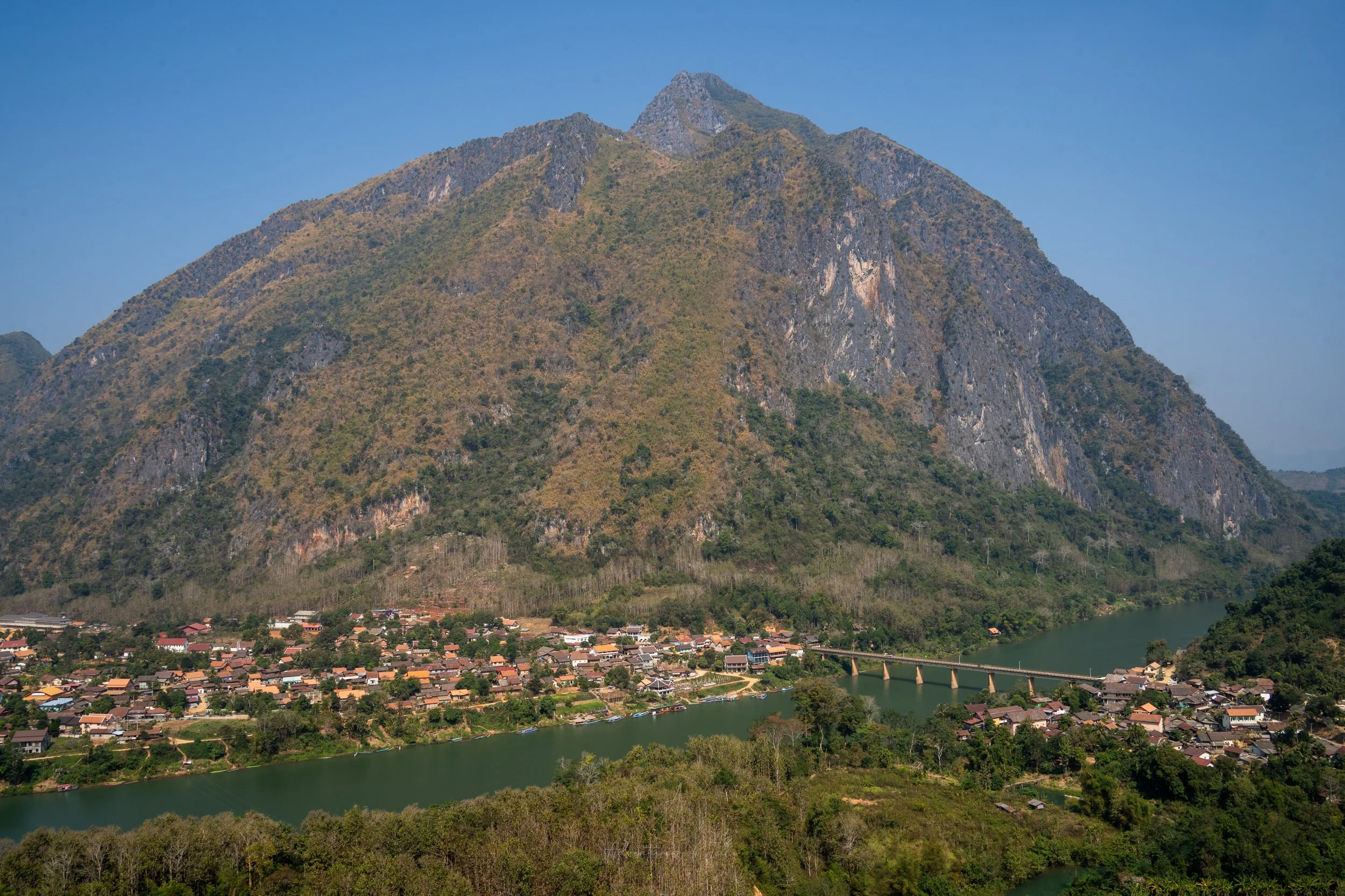 A view down over the Nam Ou river and rooftops of Nong Khiaw town in Laos. Behind the jagged, rocky, forested peak of the Sleeping Lady (Nong Khiaw’s Everest) mountain rises up to clear blue sky.