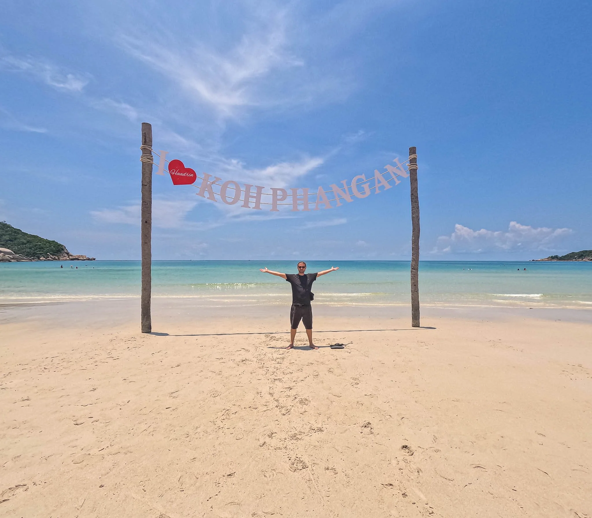 John poses for a photo underneath a sign which reads "I heart Koh Phangan". In front white sand stretches away to turquoise ocean.