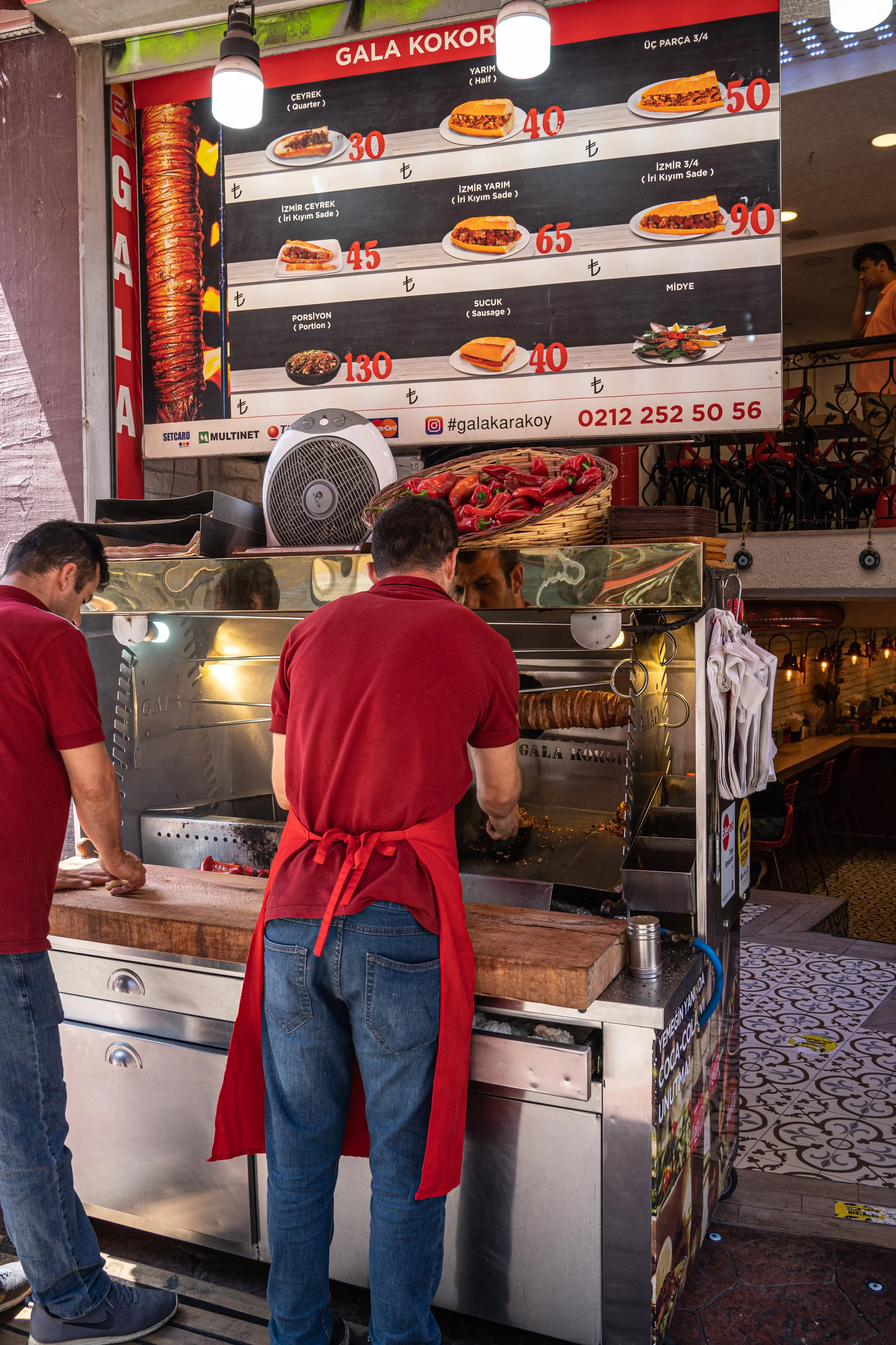 Two men wearing red t-shirts and blue jeans stand in front of a kokorec stall on the streets of Istanbul. Above them a menu displays the different korkorec on offer.