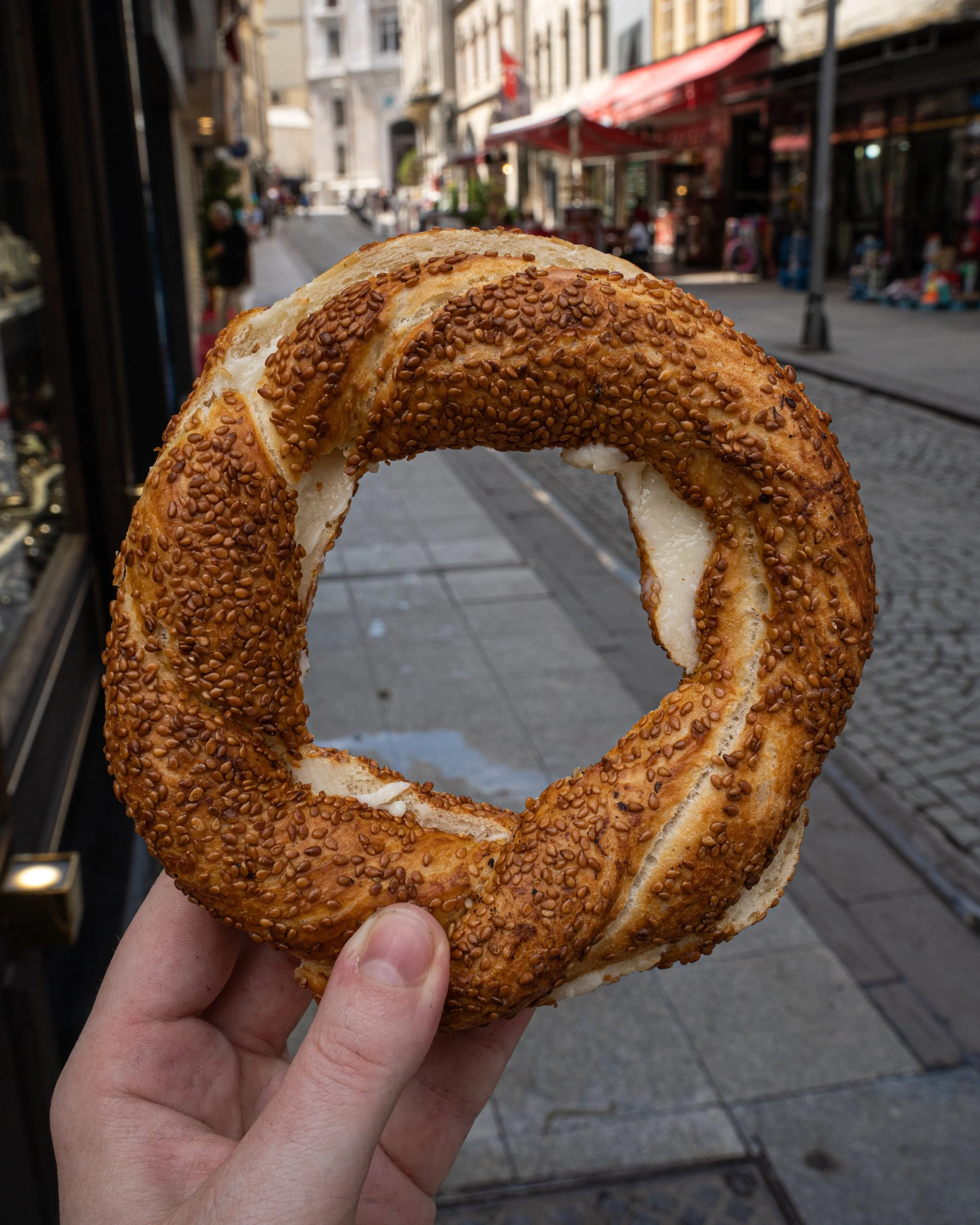 A circular simit covered in sesame seeds and stuffed with soft cheese is held up to the camera on the streets of Istanbul.