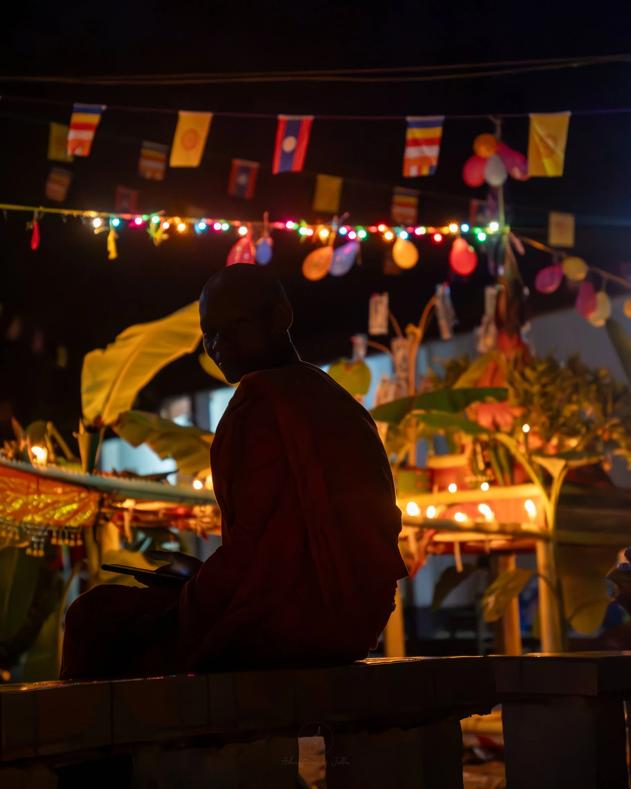 A silhouetted monk wearing orange robes looks directly into the camera during a temple festival to celebrate the end of Buddhist Lent in Nong Khiaw, Laos. Behind the monk are strings of balloons, candles and a huge wooden boat draped in offerings.