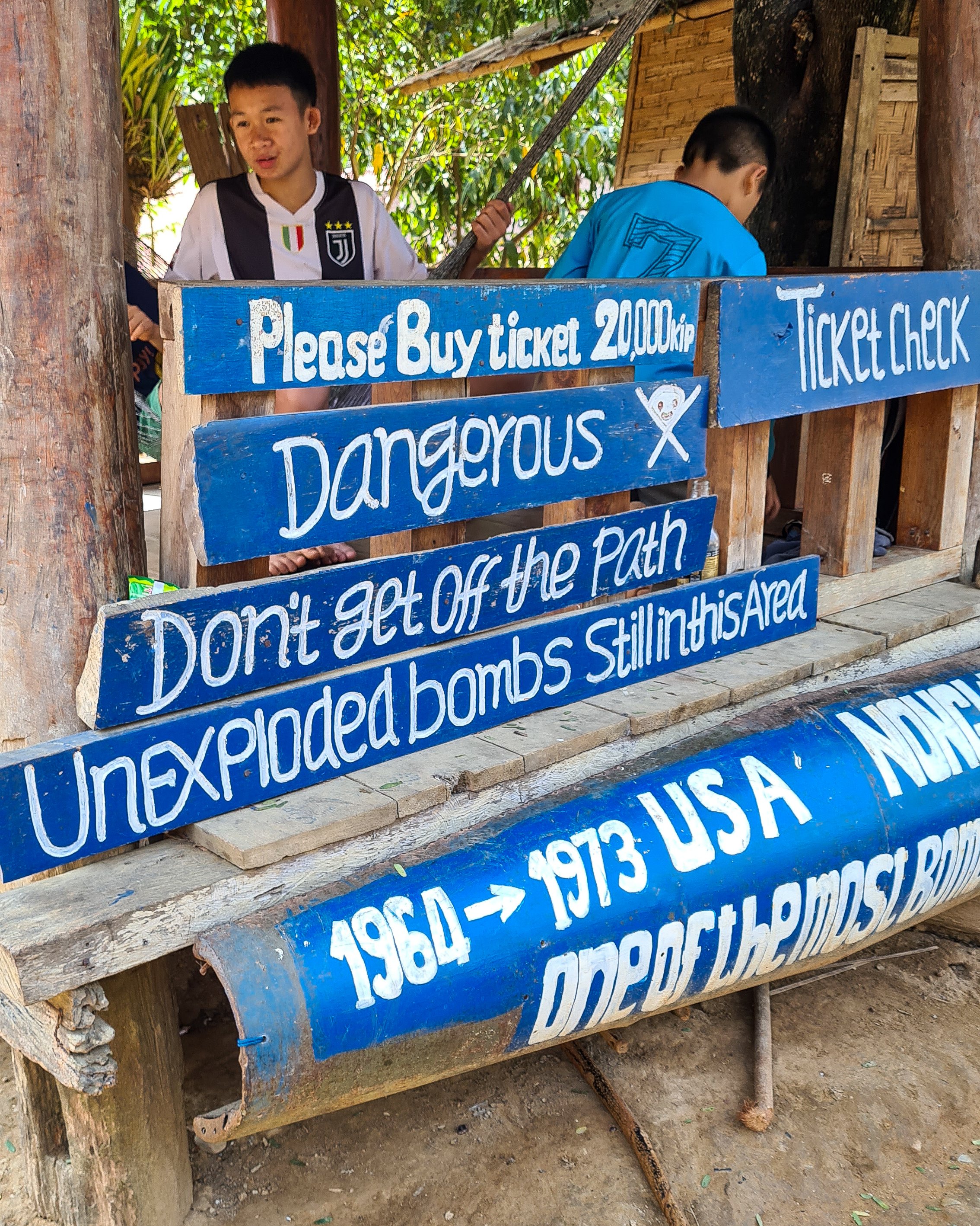 The ticket booth for the Pha Daeng hike is made up of an old bomb casing. Wooden signs are displayed reminding travellers of the dangers of unexploded bombs in Laos.