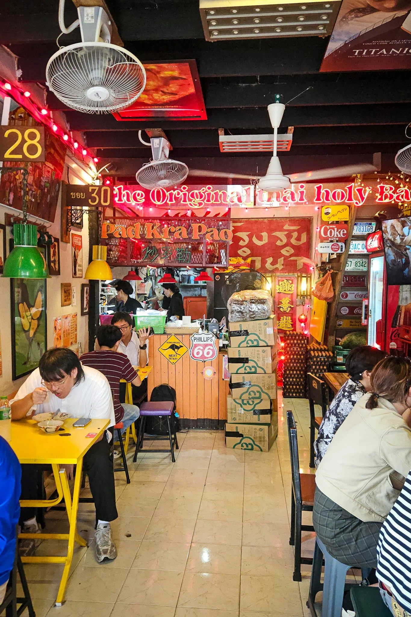 The colourful interior of Bangkok's Original Pad Kra Pao restaurant is filled with neon signs, old number plates and advertisements. The floor is filled with diners eating plates of pad kra pao.
