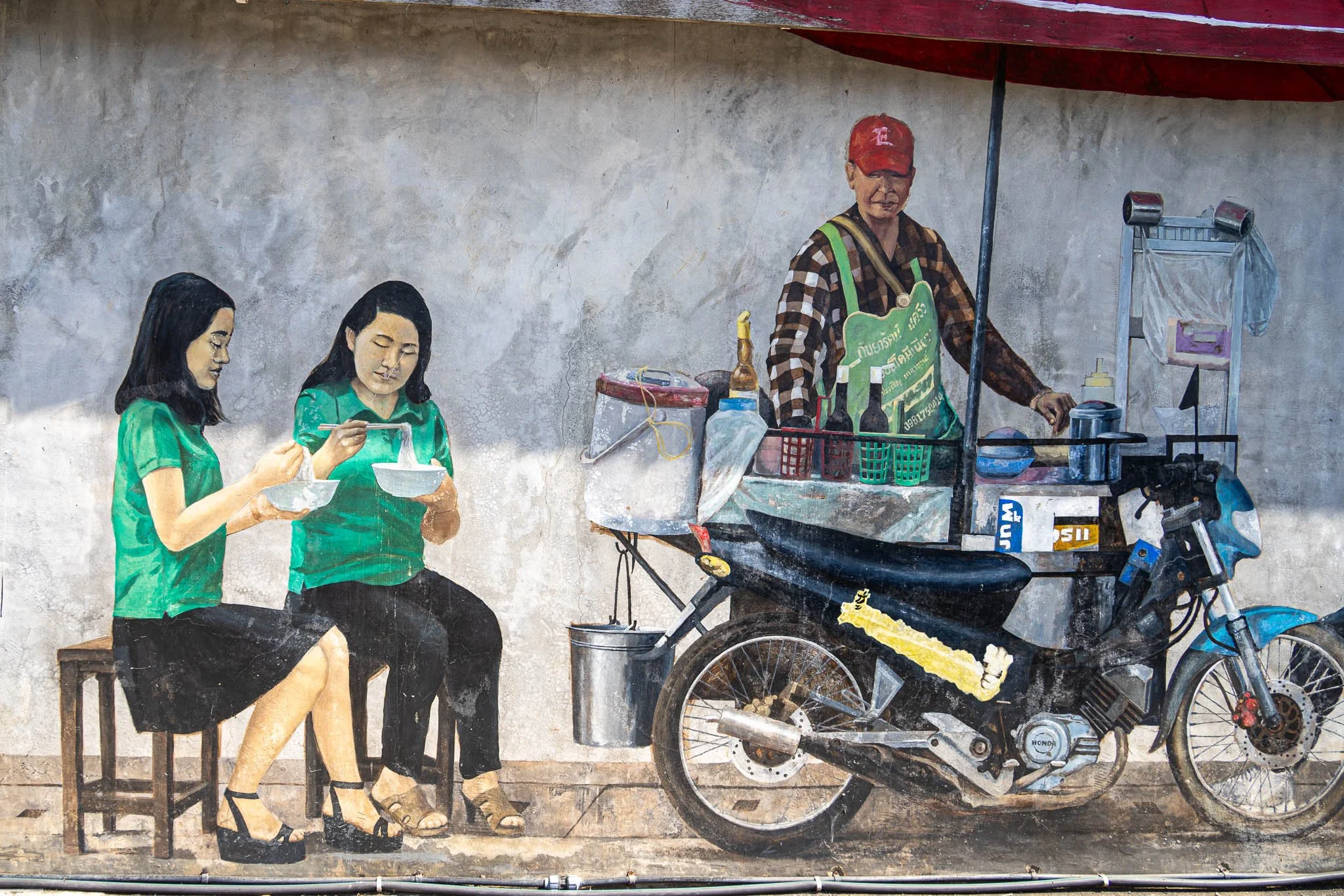 A street art mural featuring two girls slurping down a bowl of noodles next to a street food vendor is painted on a wall in the middle of Khon Kaen city, Thailand.