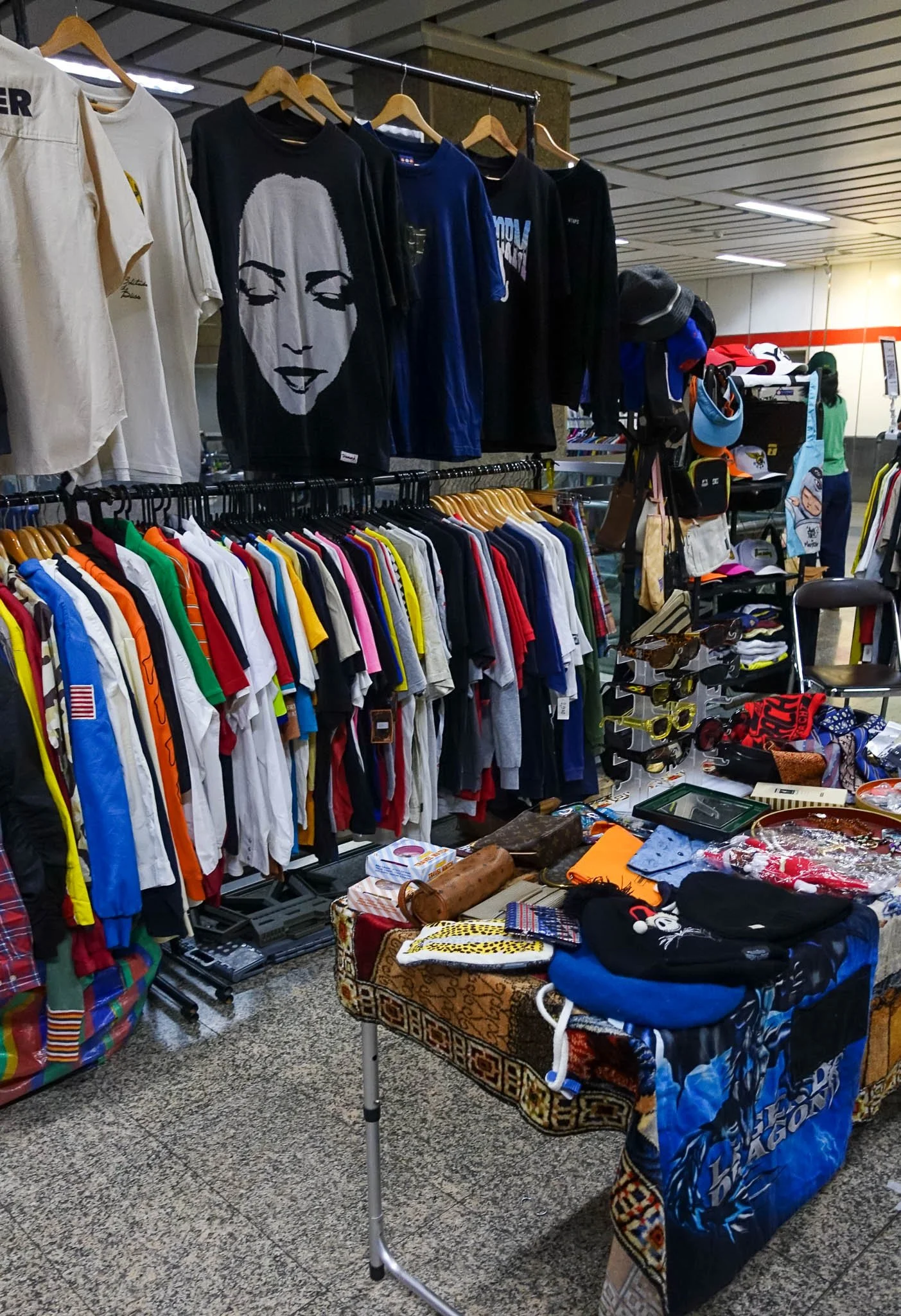 A clothes rack is full of hanging, colourful, vintage t-shirts. In front is a table full of old bags, sunglasses and scarves at the Re:Turn Market in Bangkok.