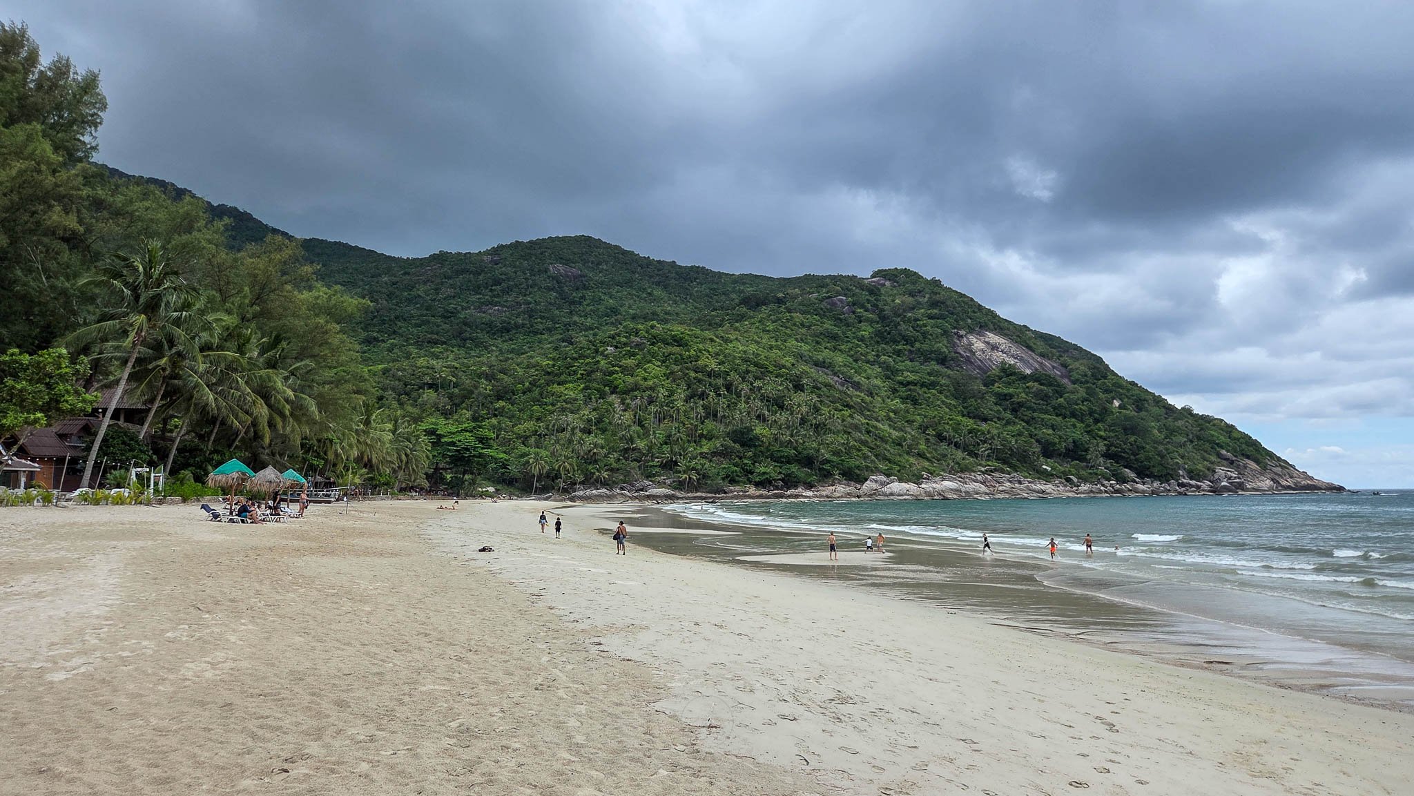 Sitting under grey clouds, the white sands of Bottle Beach stretch away from the viewer. On the beach families can be seen playing in the sea and sunbathing. Photo taken at Bottle Beach on Koh Phangan.