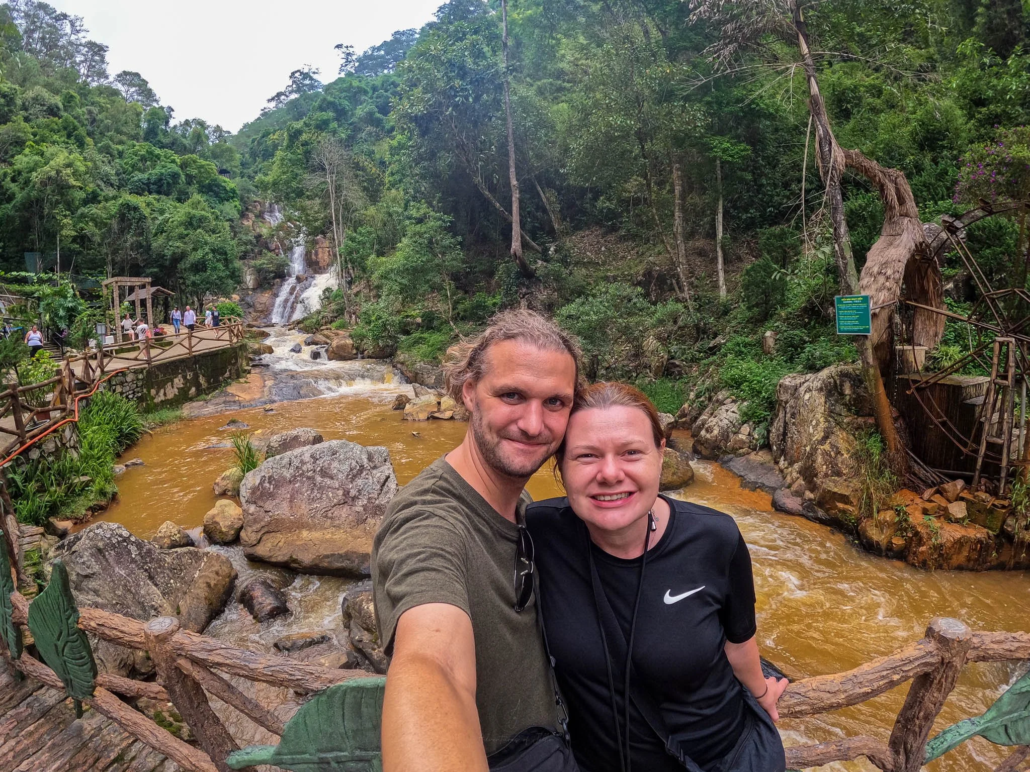 Adventures of Jellie smile at the camera. Behind them the white waters of the Datanla Waterfall crashes down into a brown river.