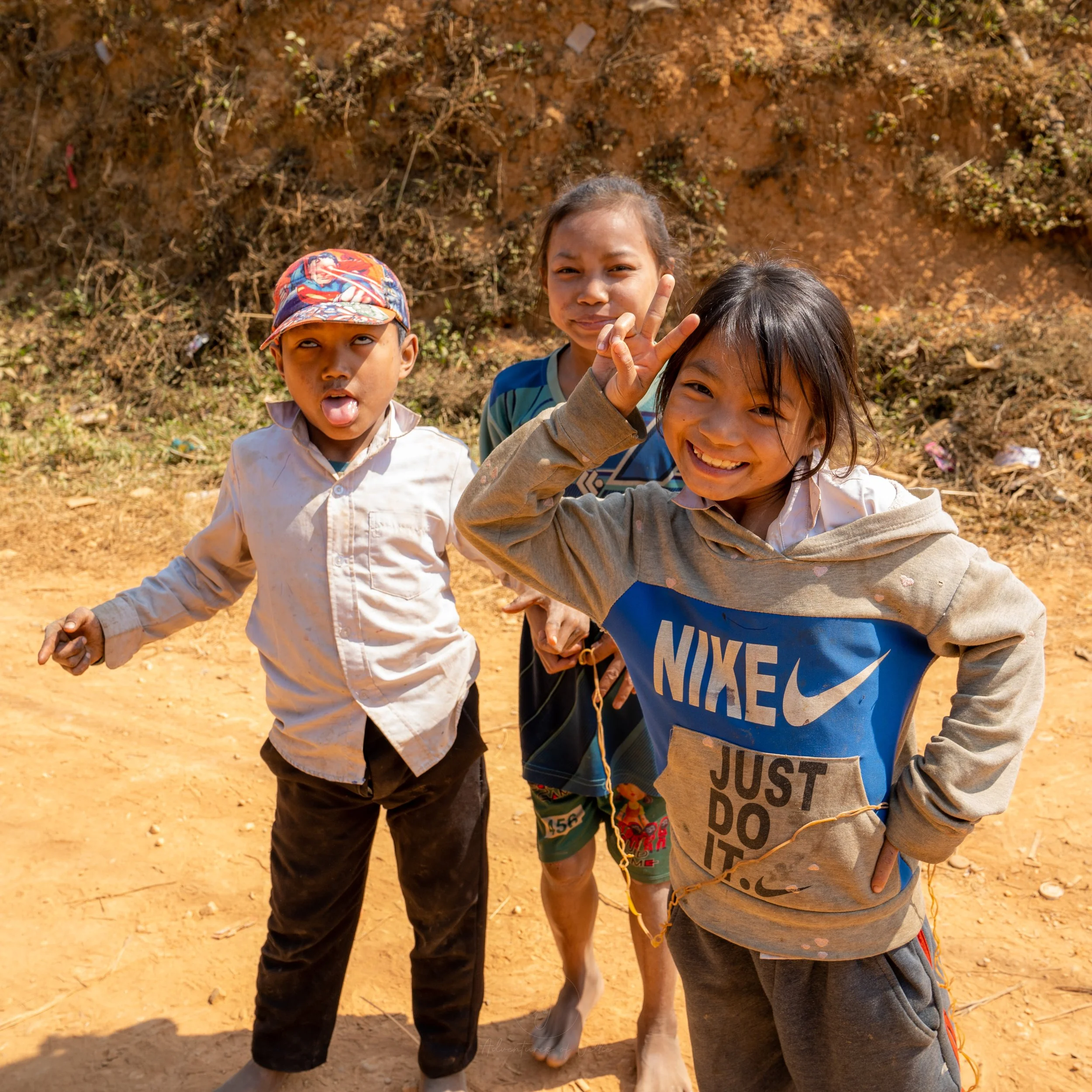 A group of children pose for a photo whilst making silly faces. The boy to the left is sticking his tongue out, the girl in the middle is smiling and the girl to the left holds up a peace sign in a village outside of Nong Khiaw in Laos.