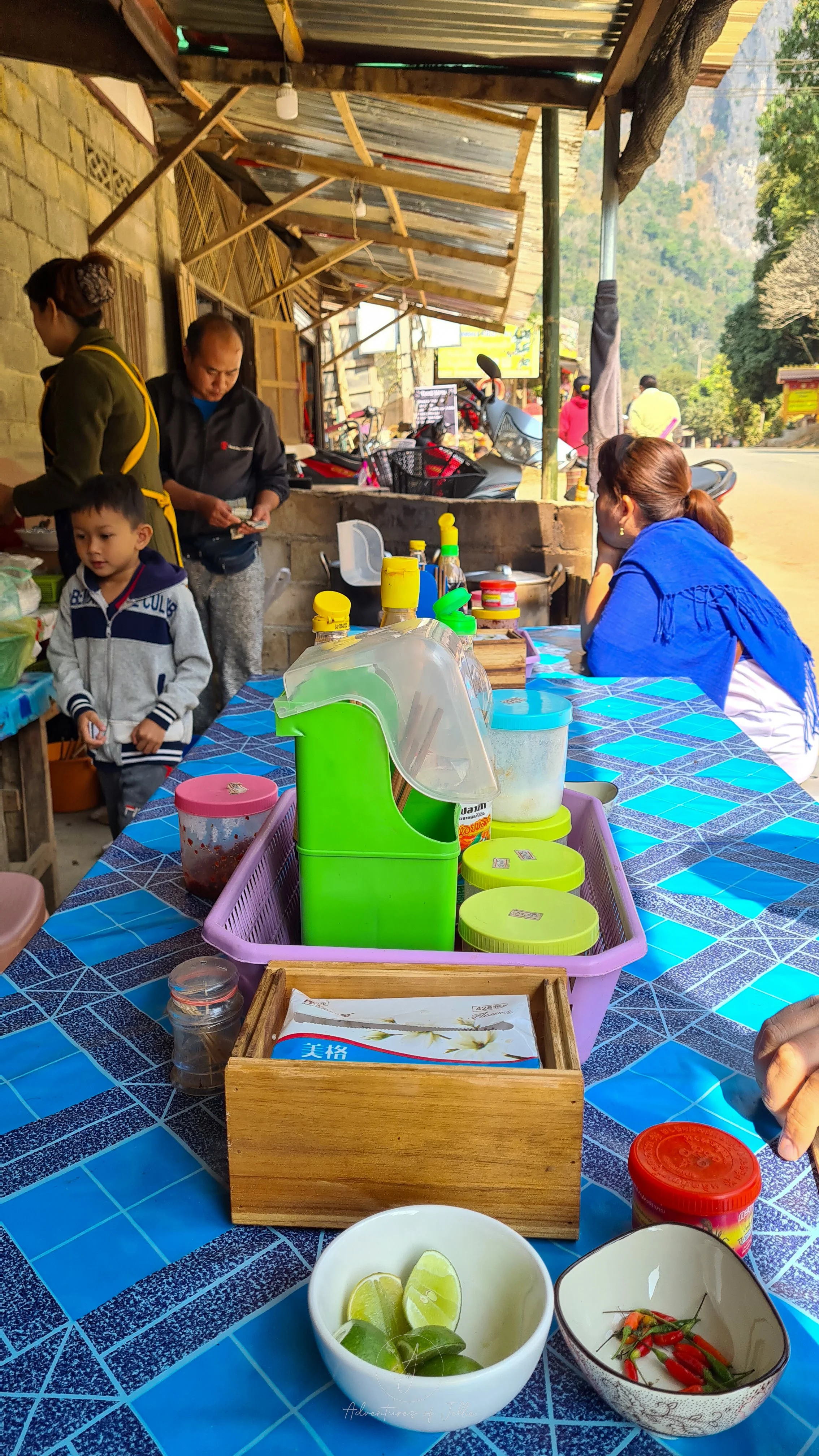 Locals from Nong Khiaw in Laos sit around a table at a street side noodle soup stall. On the blue table are plastic containers in various sizes filled with chopsticks, spoons and condiments.