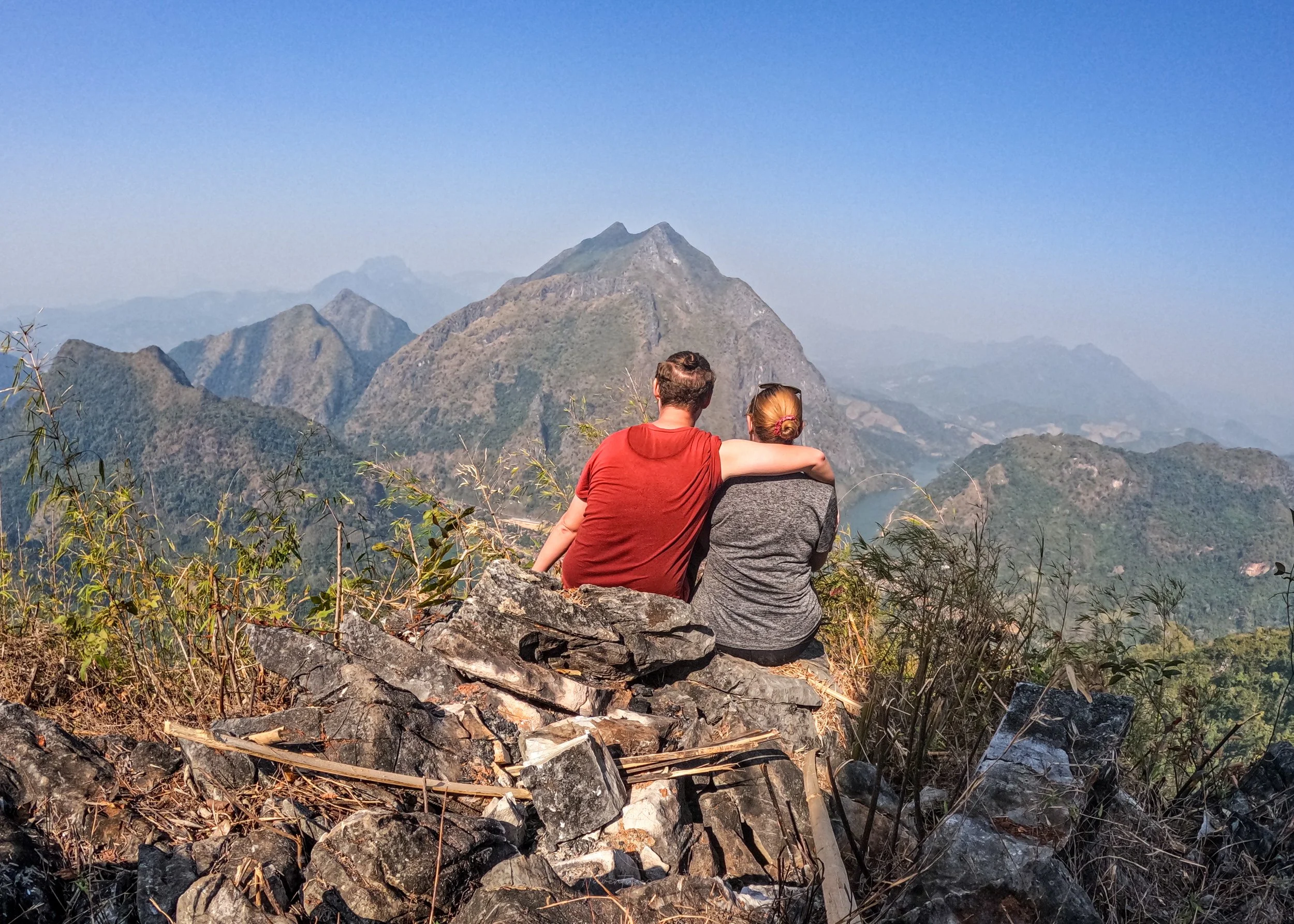 Adventures of Jellie sit on a rock with their arms around each overlooking the town and mountains of Nong Khiaw. Photo taken from the viewpoint at the top of the hiking trail on Pha Kao mountain.