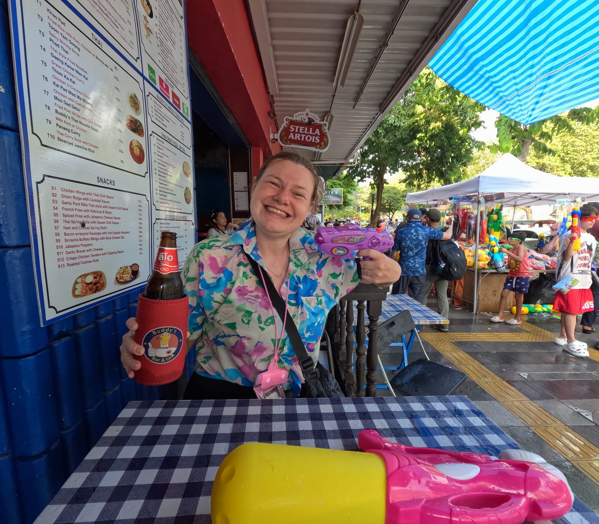 Ellie sits at a table with a large Leo beer, soaked but grinning from ear to ear. She is gesturing to the beer with a purple My Little Pony water pistol.
