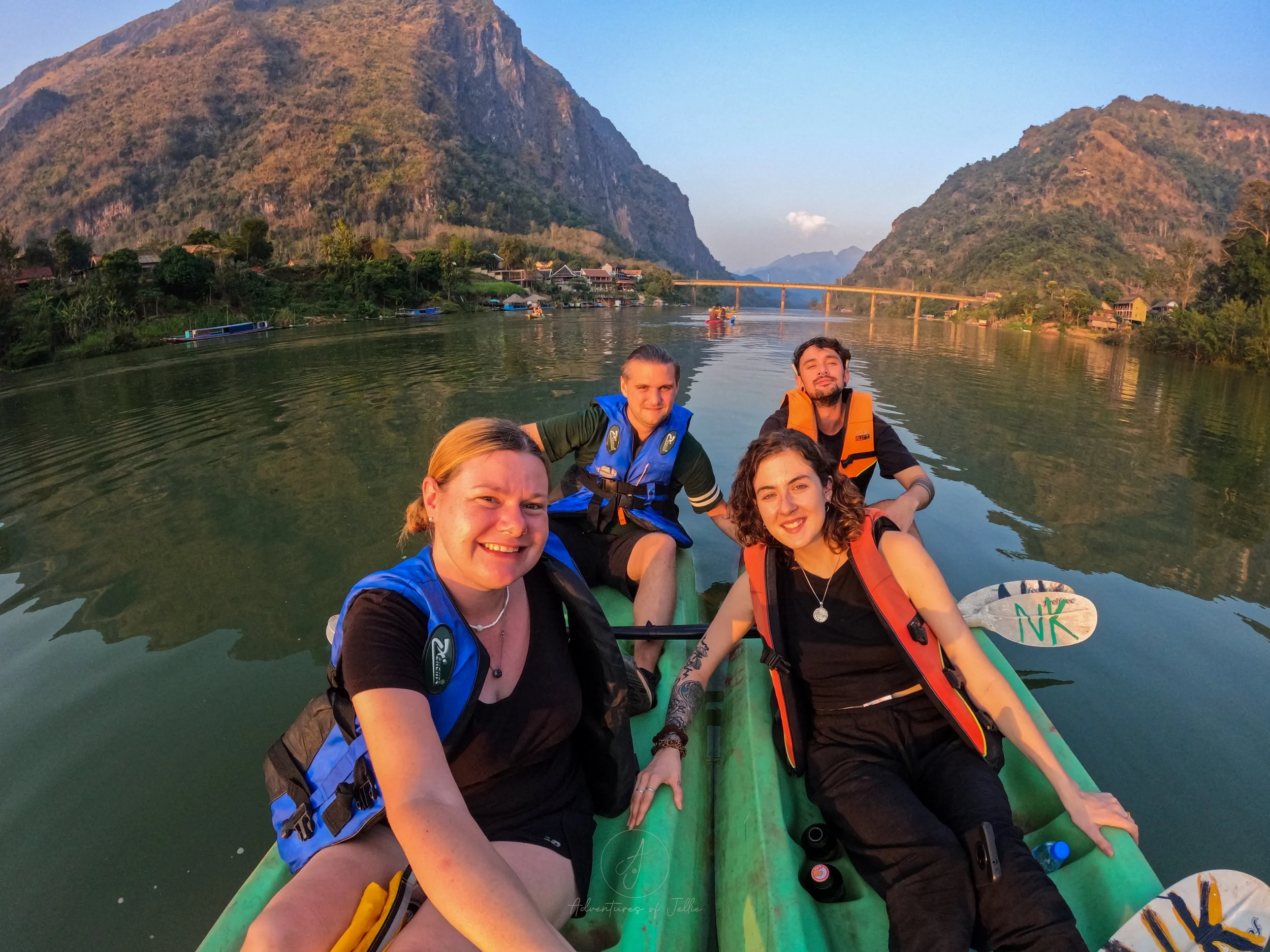 Adventures of Jellie sit with friends whilst floating on two green kayaks in the middle of the Nam Ou River in Nong Khiaw. The whole group is bathed in a warm glow from the setting sun.