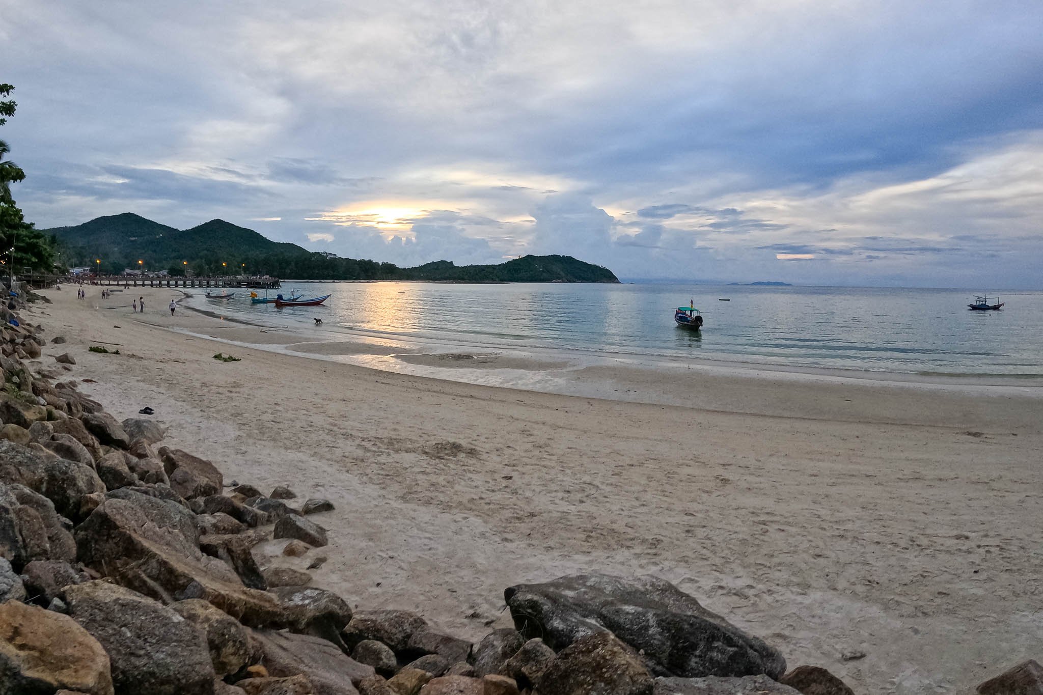 The sun sets over Chaloklum Beach. In the bay fishing and longtail boats sit moored under the grey sky.
