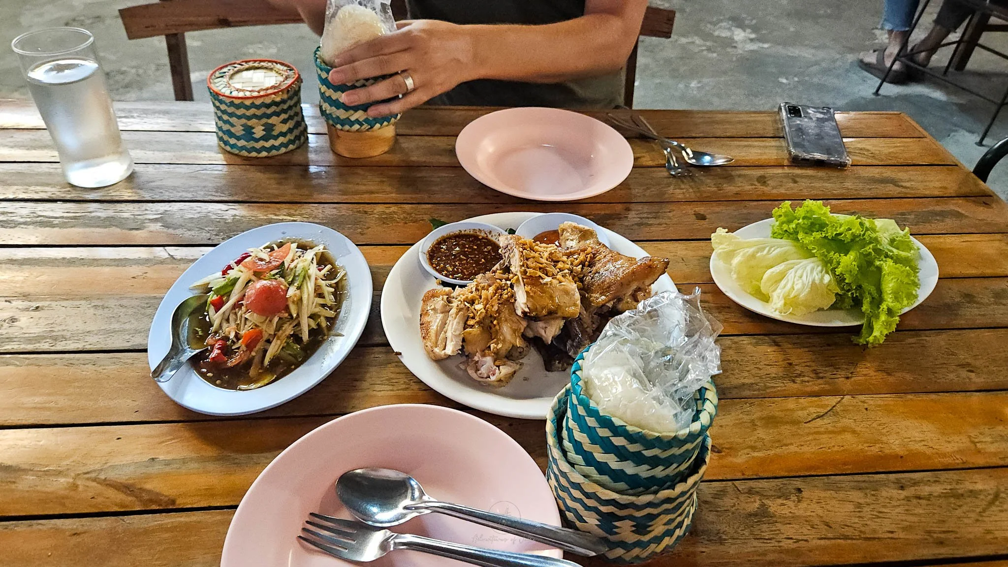 On a wooden table sits a plate piled high with chunks of grilled chicken. Next to this is a plate of sliced som tam salad and a plate of salad leaves. In front of the chicken is a basket of sticky rice.