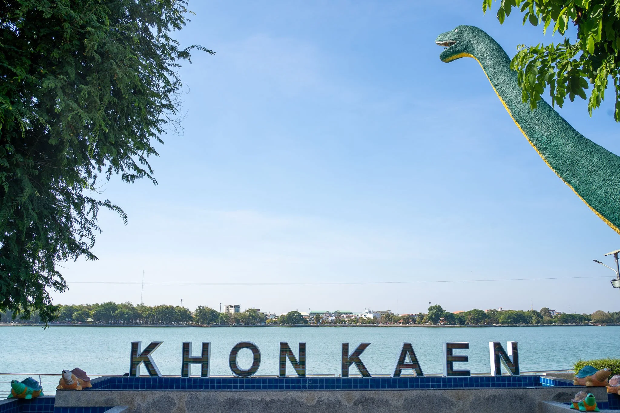 A green diplodocus statues towers over a blue lake. In the foreground a sign written in capital letters reads "Khon Kaen".